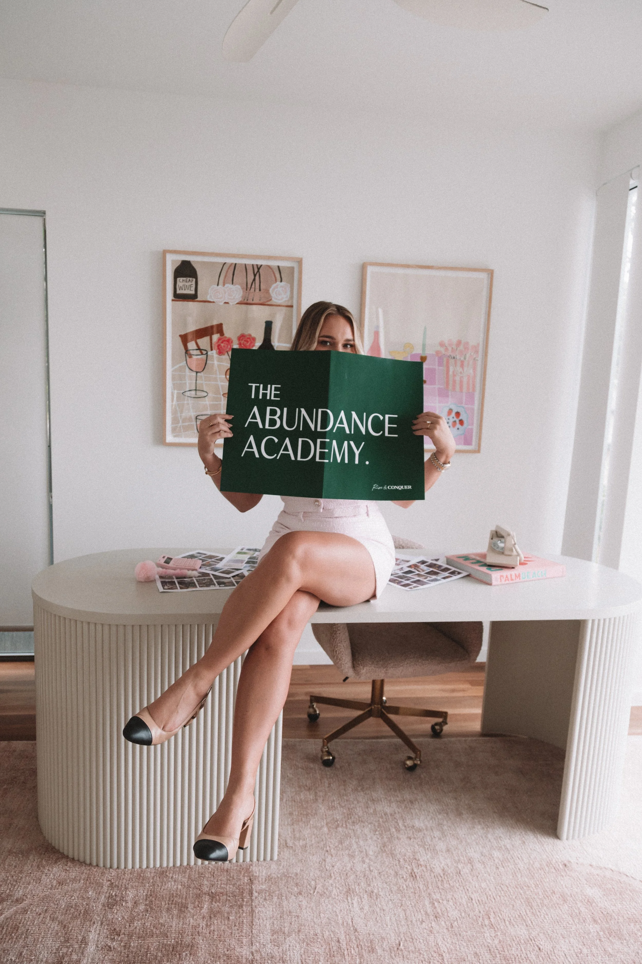 A woman sitting at a white curved desk, holding a green sign that reads "The Abundance Academy." She is wearing a pink skirt and black-and-beige shoes, with her crossed legs resting on the desk. Behind her, there are two framed art pieces on a white wall, and a pink book on the desk titled "Palm Beach." There is also a vintage phone and a pile of photographs on the desk, and a beige office chair with caster wheels in the background.
