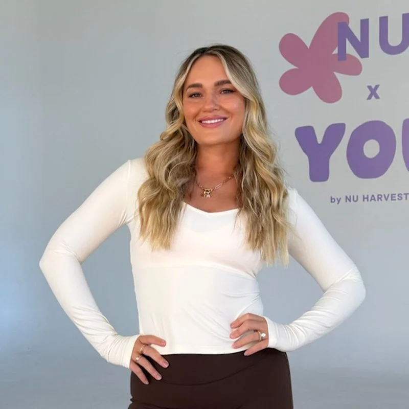 A smiling woman with long, wavy blonde hair wearing a white long-sleeve top and black pants, standing in front of a purple and pink logo with the words 'NU x YOU' and 'by NU HARVEST' on a light-colored wall.
