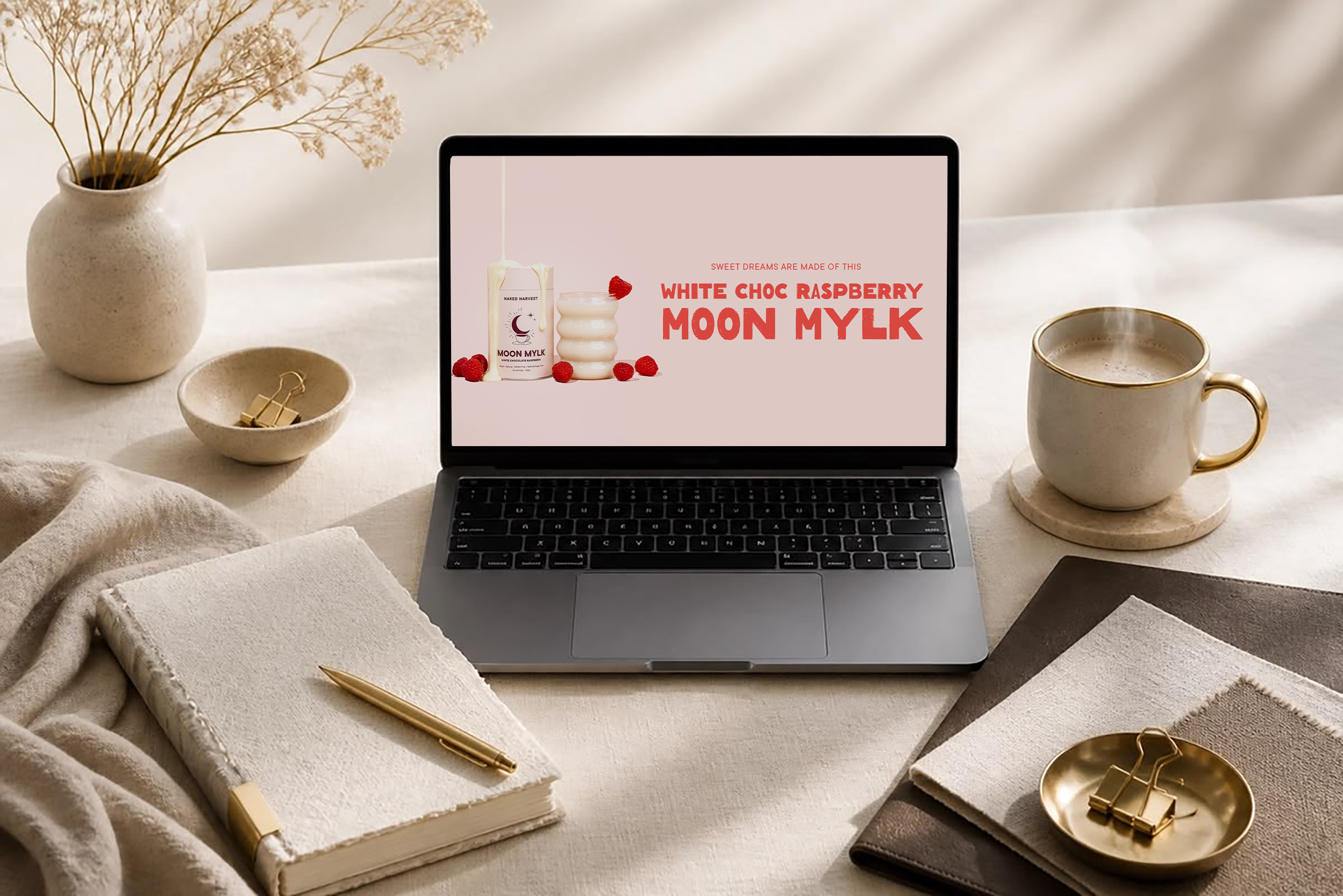 A laptop displaying an advertisement for a product called 'White Choc Raspberry Moon Mylk' on a desk, surrounded by a notebook, a gold pen, a cup of coffee, decorative dishes with gold clips, and a beige vase with dried white flowers.