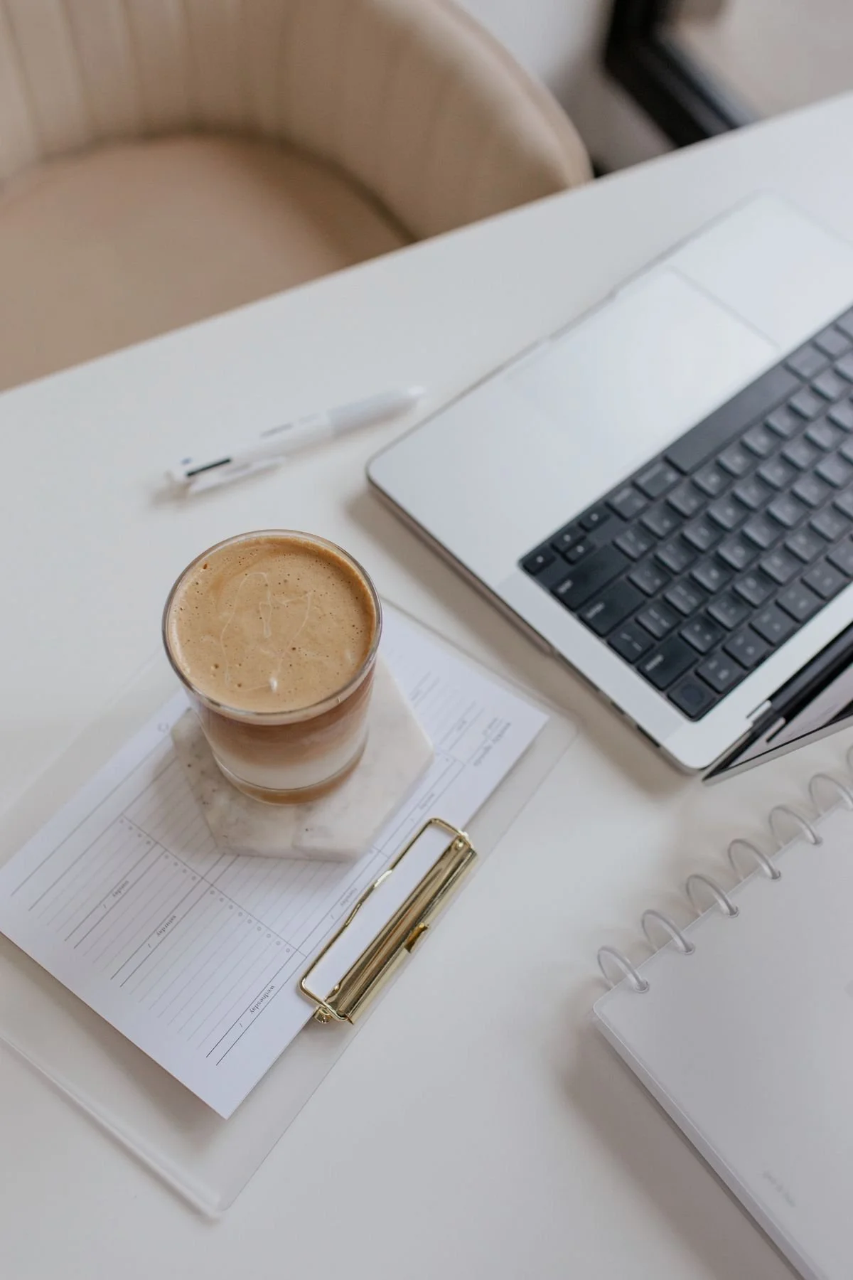 Workspace with a laptop, coffee, notepad, clipboard, and a pen on a white desk.