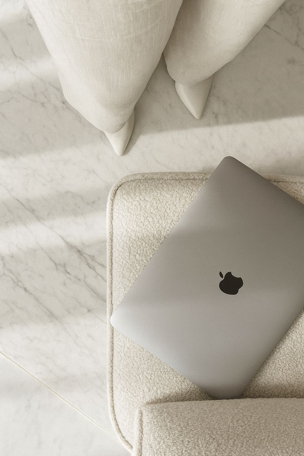 A closed silver Apple MacBook laptop resting on a beige textured armrest of a sofa, with a white marble floor and part of a white curtain in the background.