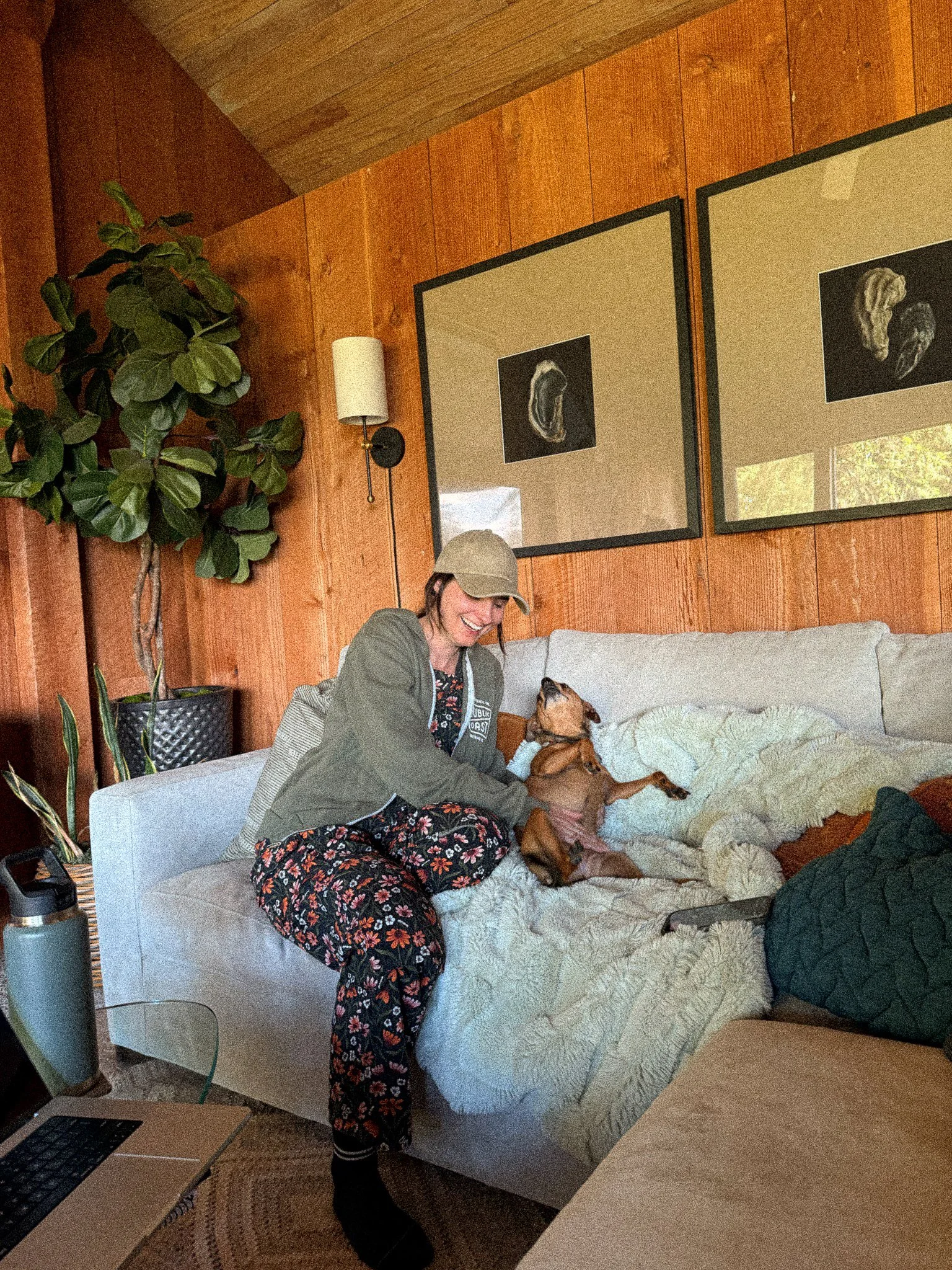 A woman sitting on a bed, smiling and looking at a dog lying on the bed. The room has wood-paneled walls, artwork hanging, a large potted plant, and a lamp. A laptop and a water bottle are on a nearby table.