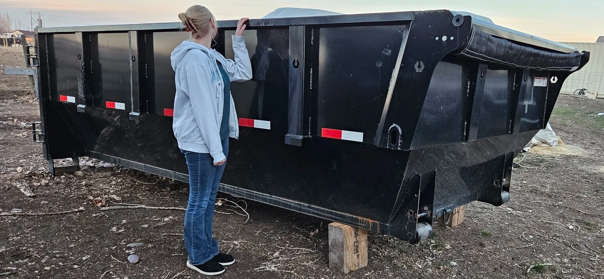 A person with blonde hair in a ponytail, wearing a light gray hoodie, blue jeans, and black shoes, stands next to a large black dumpster on a dirt lot, looking at it with their hand on its edge.