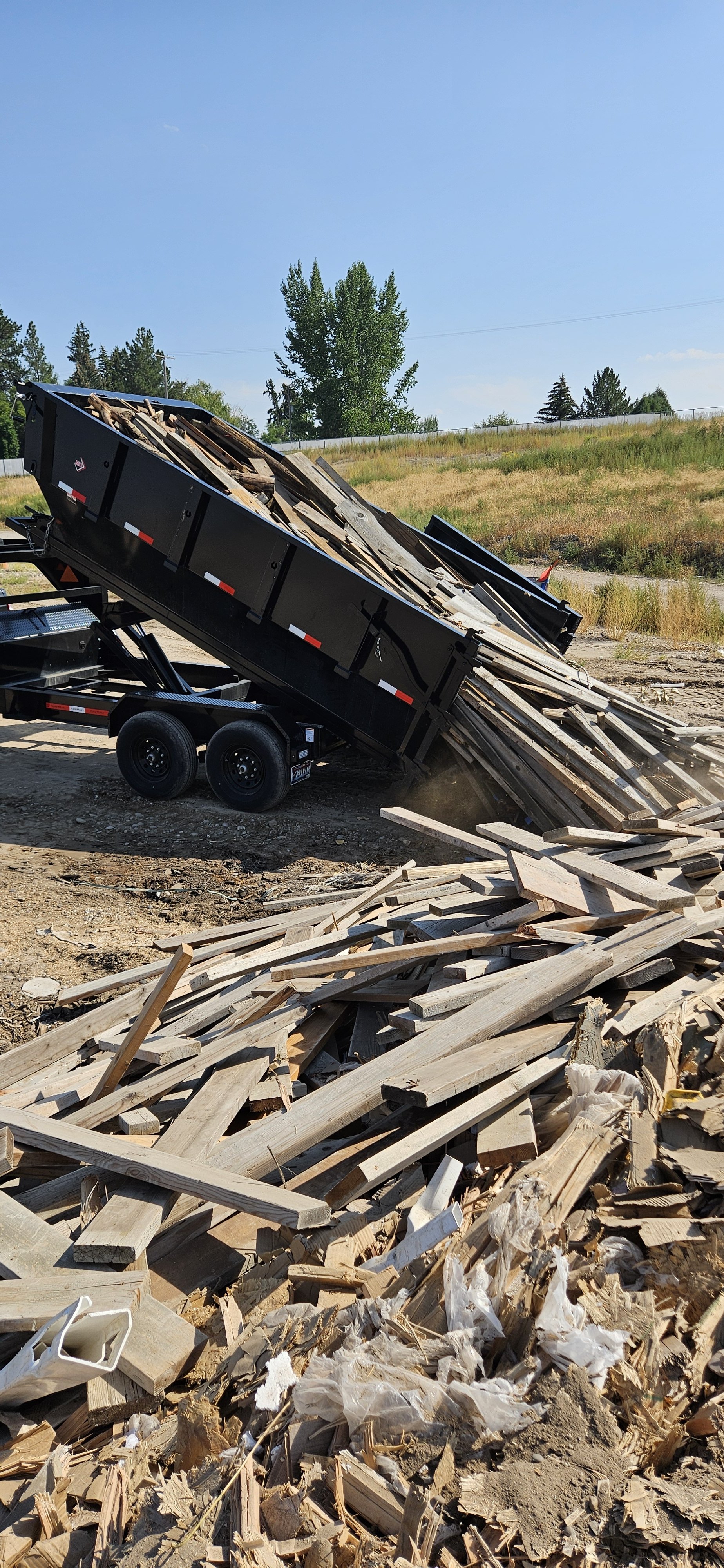 A black flatbed trailer tilted, unloading wooden debris and scraps onto the ground at a construction or demolition site.