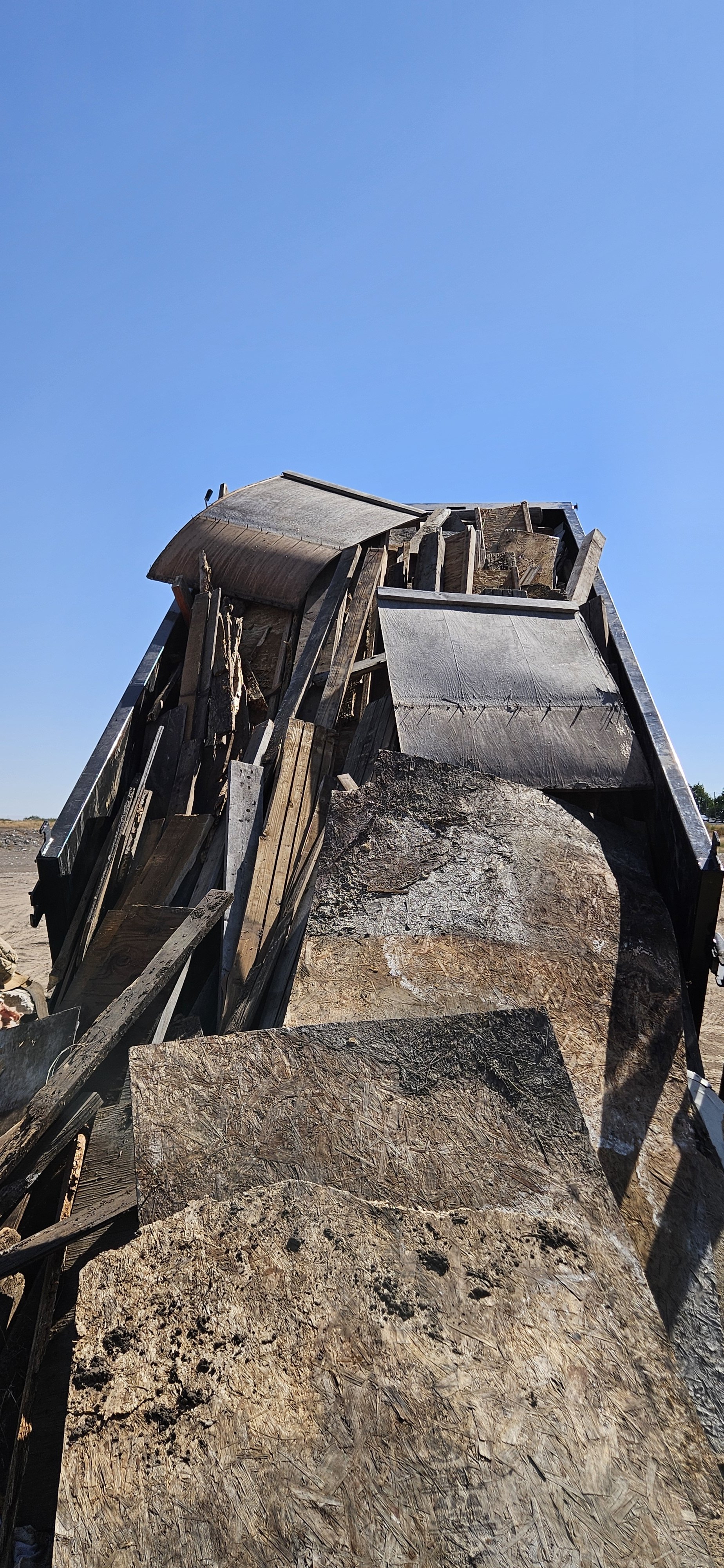 A pile of broken, weathered wood and debris standing upright outdoors against a clear blue sky.