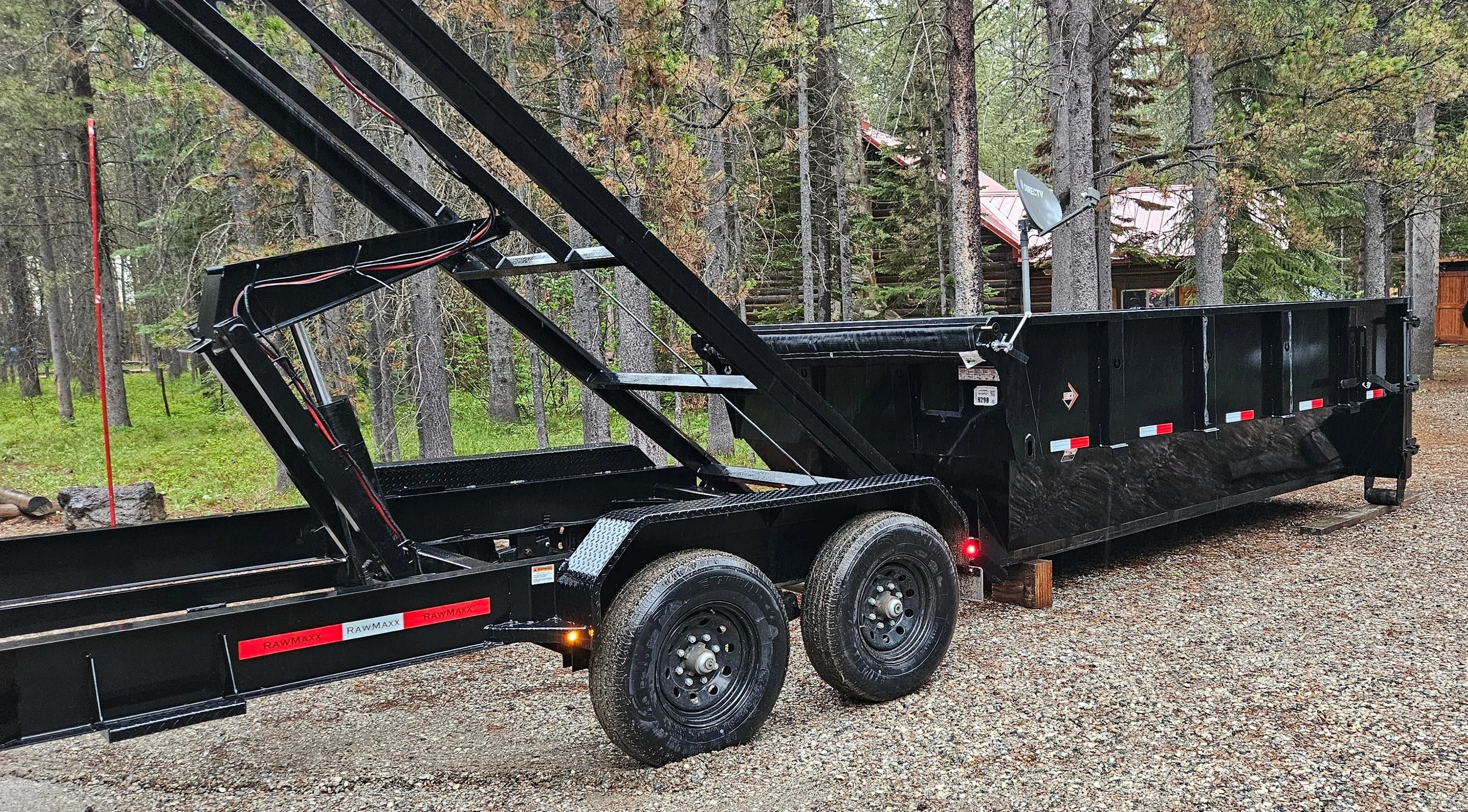 A black utility trailer with a hydraulic hydraulic lift system parked on a gravel driveway surrounded by trees and a wooded area in the background.