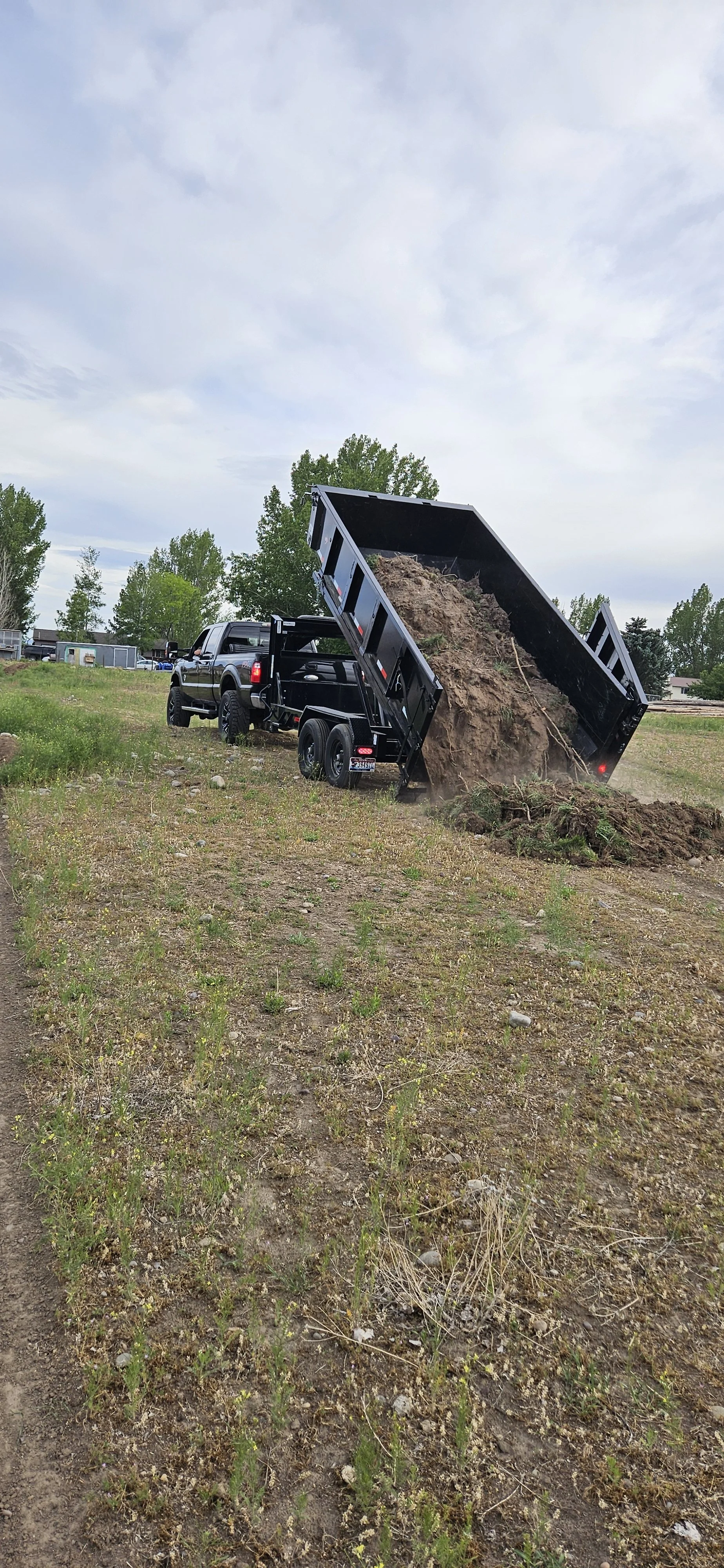 A black dump truck unloading dirt or soil onto a patch of dry, grassy land in an open field with trees and cloudy sky in the background.