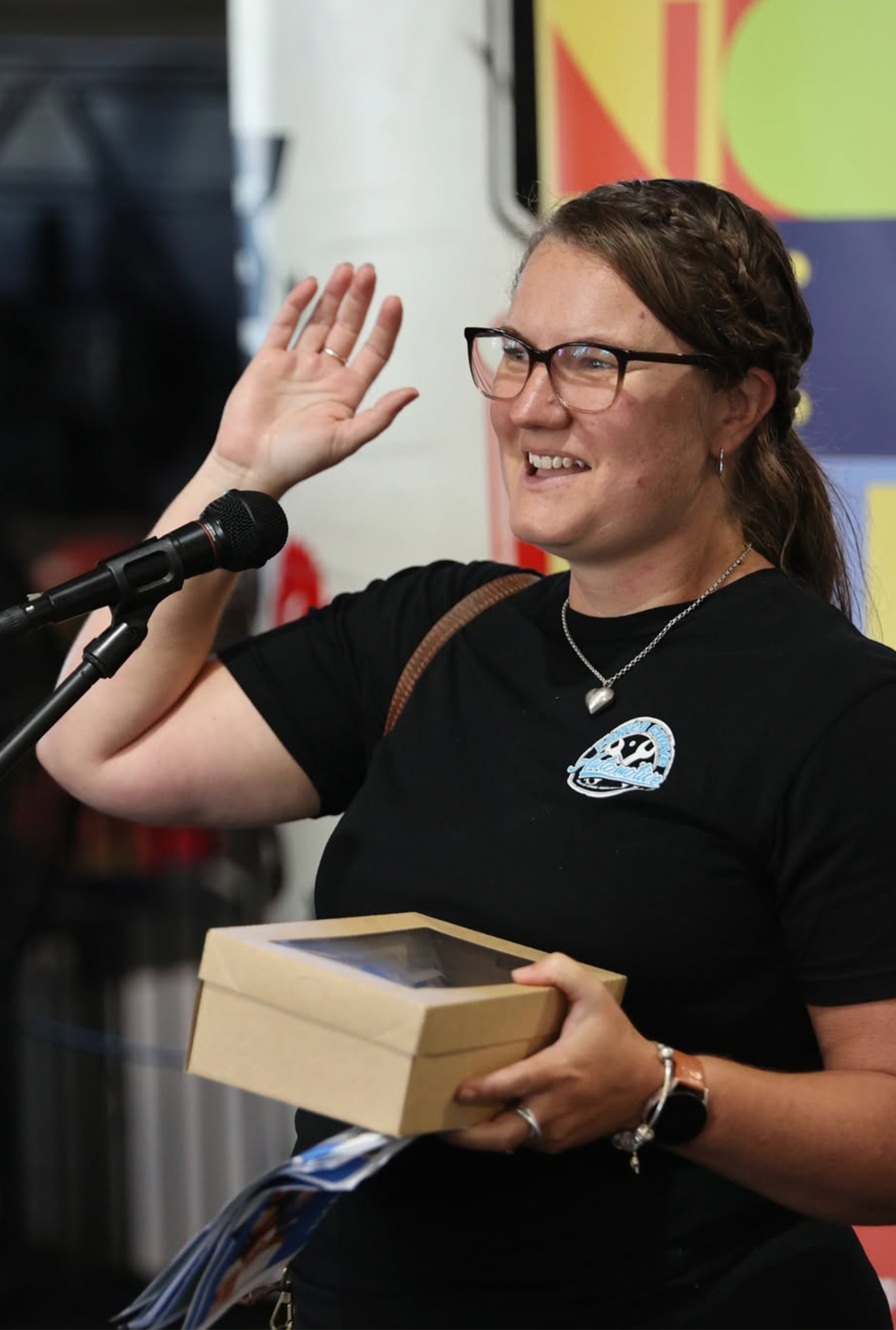 A woman wearing glasses, a black t-shirt with a logo, and jewelry, smiling and holding a cardboard box, with her right hand raised in a wave.