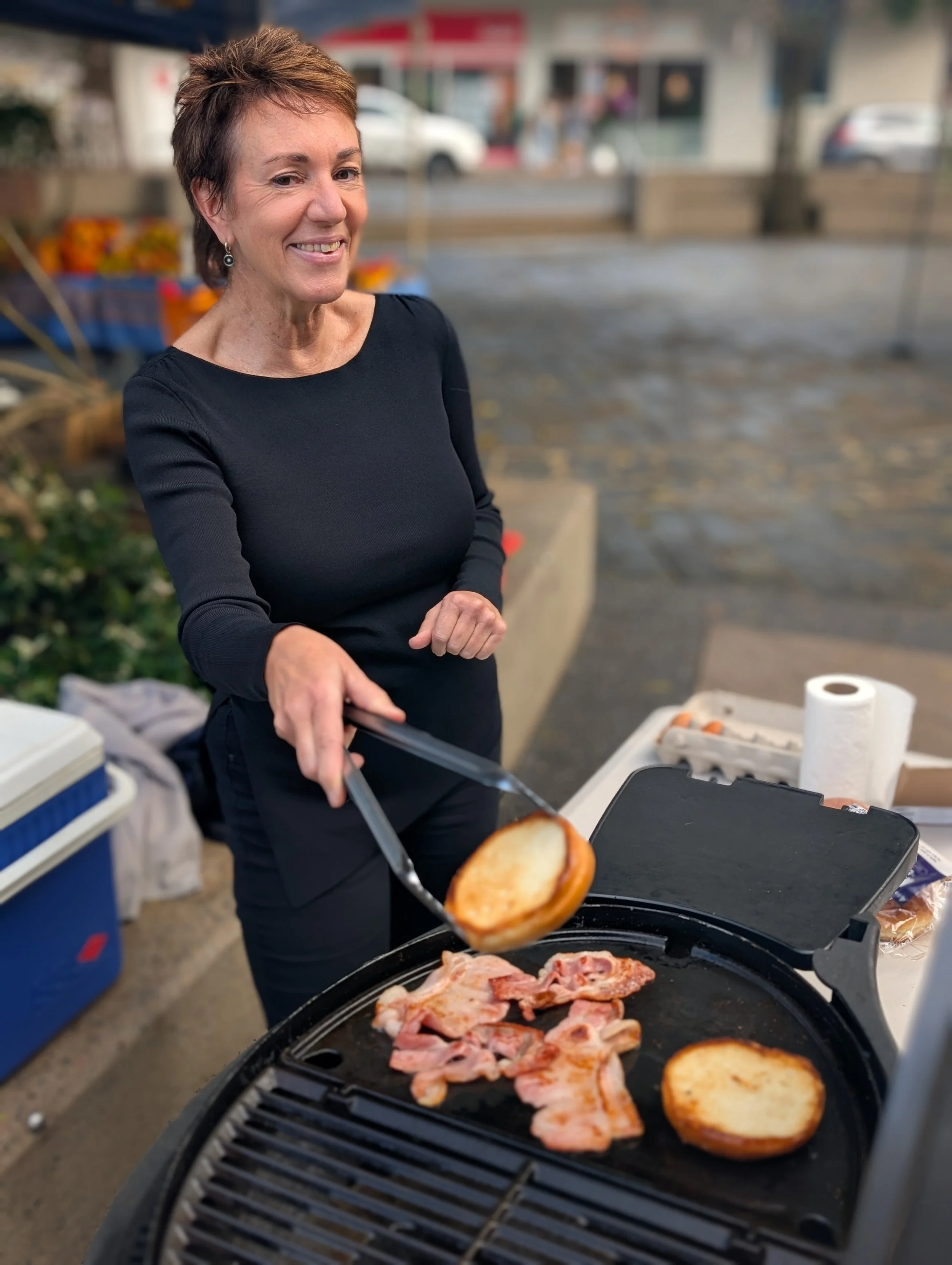 A woman grilling bacon and bread on an outdoor grill, with a blurred parking lot and storefronts in the background.