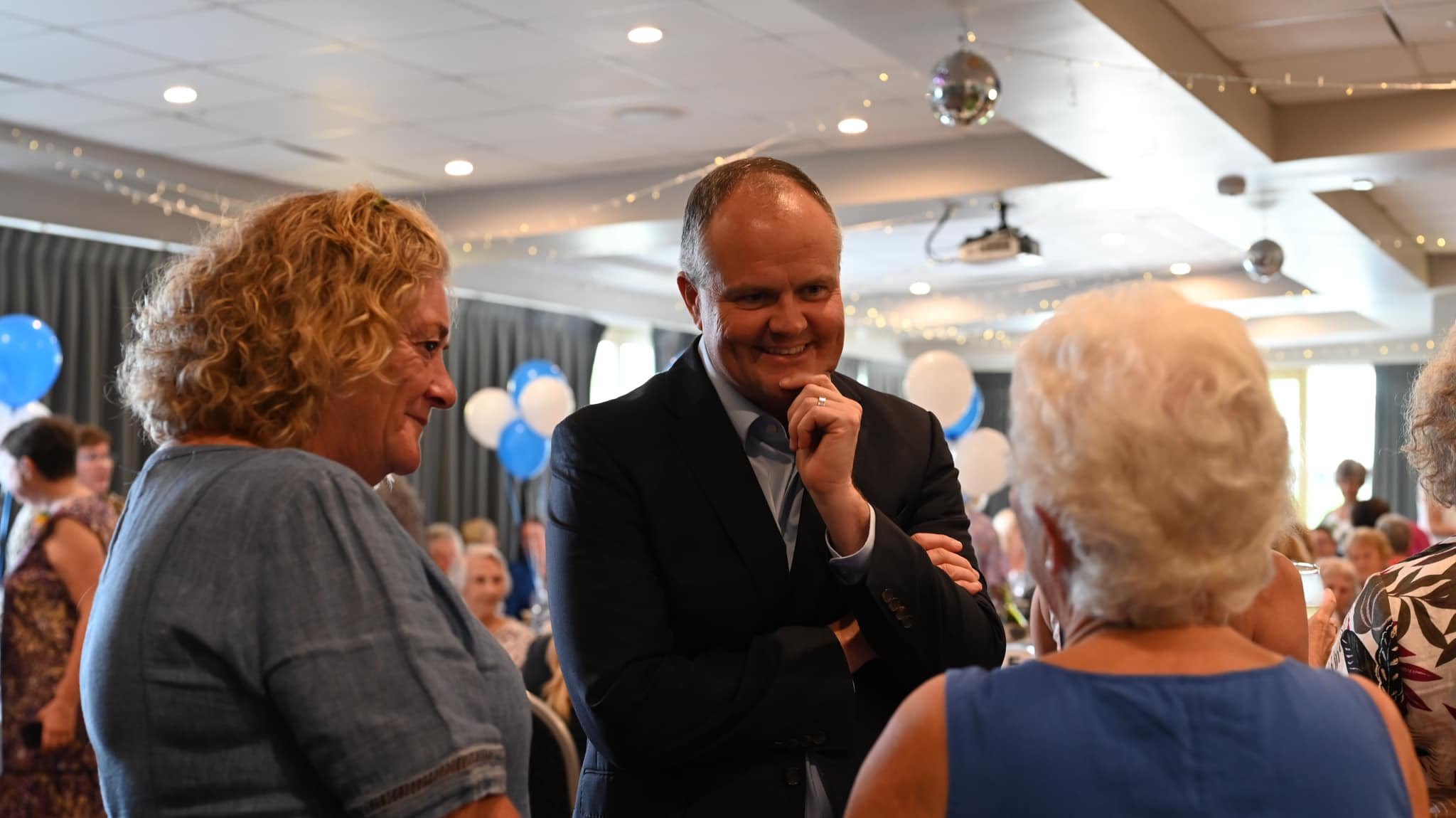 People attending a social event, engaging in conversation in a decorated banquet hall with blue and white balloons and fairy lights.