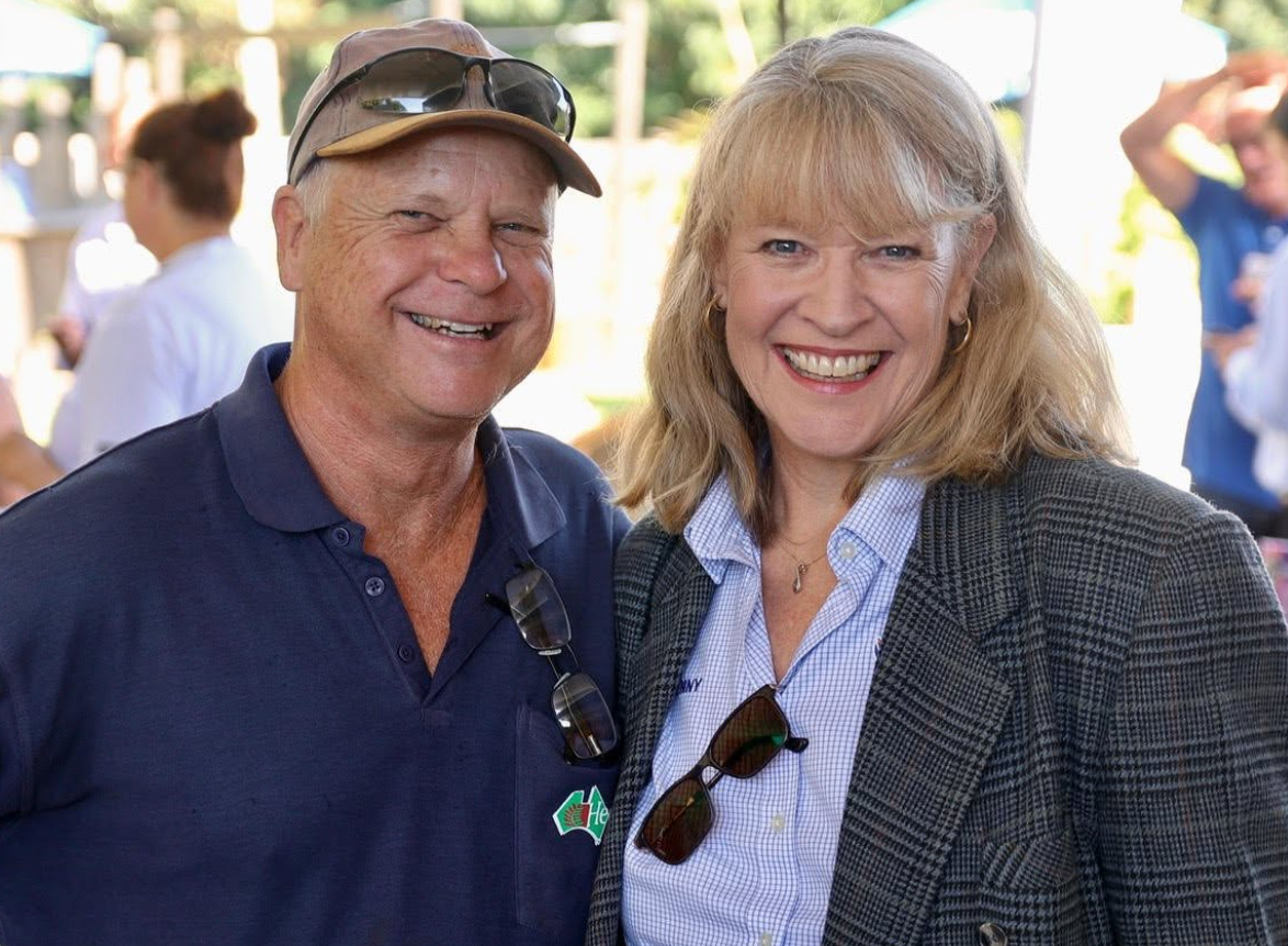 A man and a woman smiling at an outdoor event, with other people in the background.