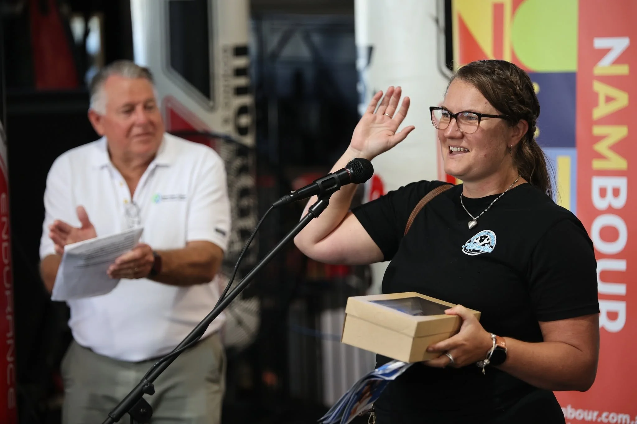 A woman with glasses and a black T-shirt is speaking into a microphone while holding a small cardboard box. There is a man in a white shirt with a paper in the background, reaching toward the woman and looking at her. They are at an indoor event with a brightly colored banner in the background.