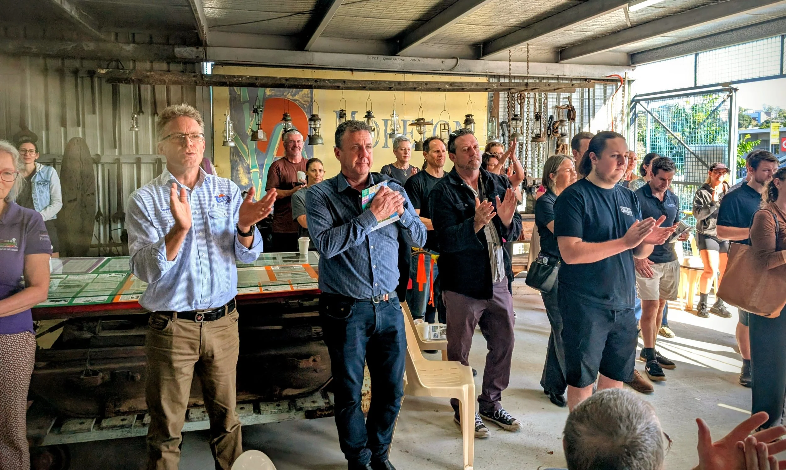 People attending an event inside a rustic building with hanging lamps and a large yellow sign in the background, some clapping and listening attentively.