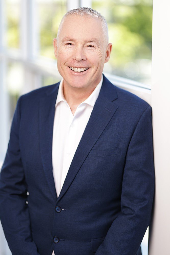 A smiling middle-aged man with short gray hair, wearing a dark blue suit and white shirt, standing next to a window with a green outdoor scene in the background.