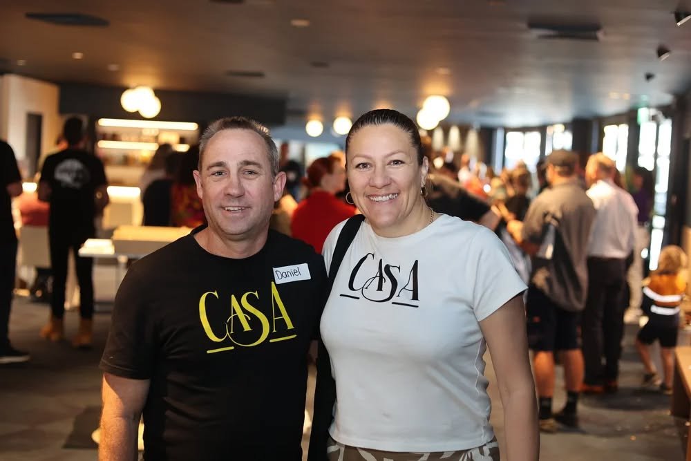 A man and woman smiling at an indoor event, wearing matching 'CASA' t-shirts, with other people in the background.
