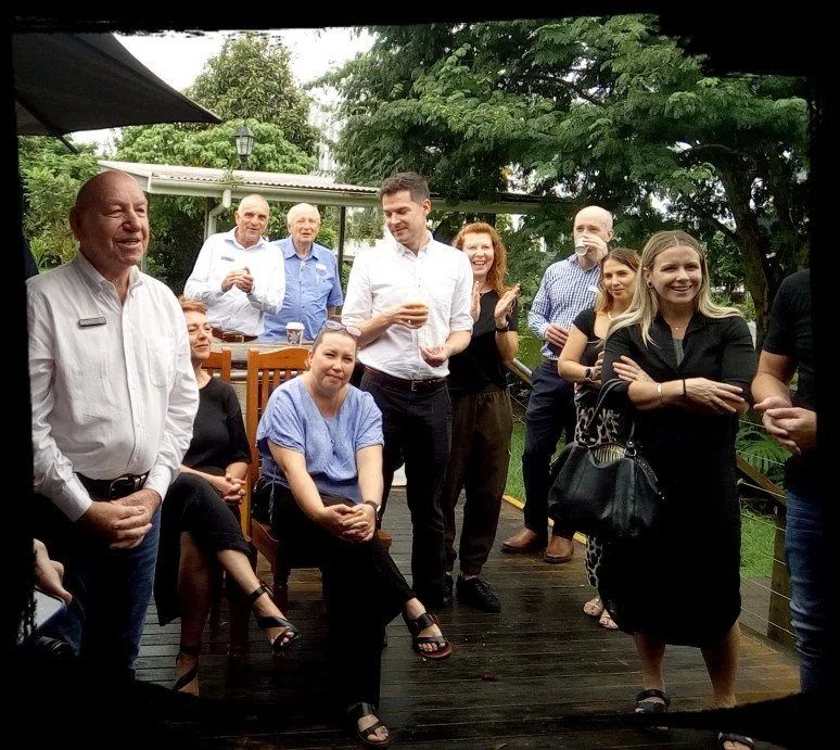 Group of people gathered outdoors on a wooden deck, smiling and engaged in conversation, with lush green trees in the background.