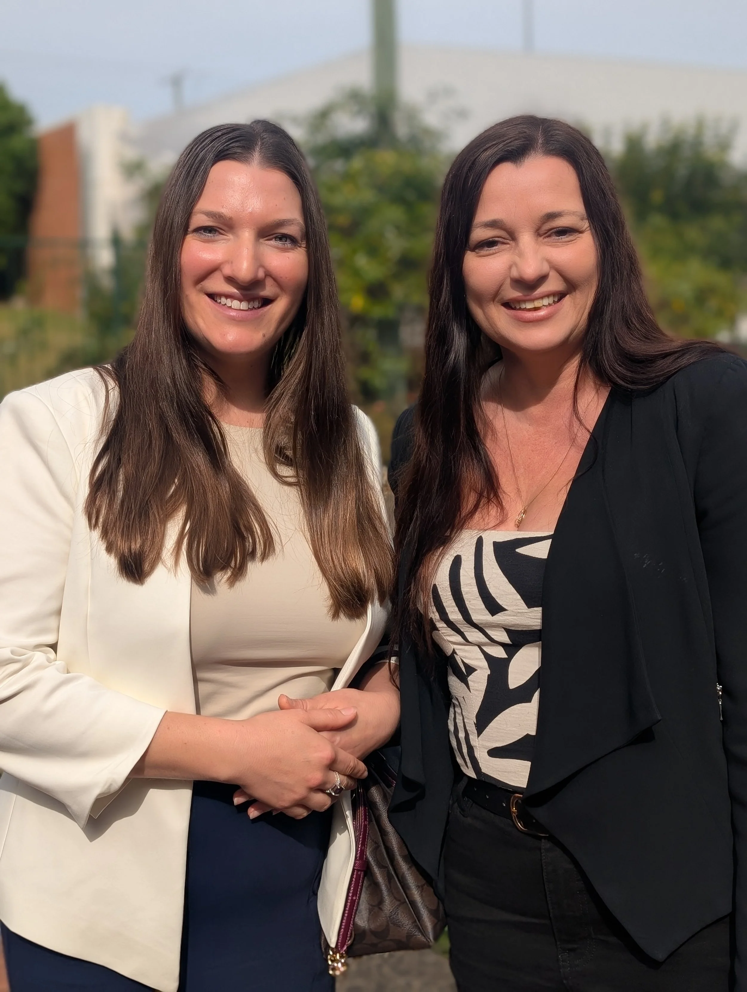 Two women with long dark hair smiling and holding hands outdoors, with trees and a building in the background.