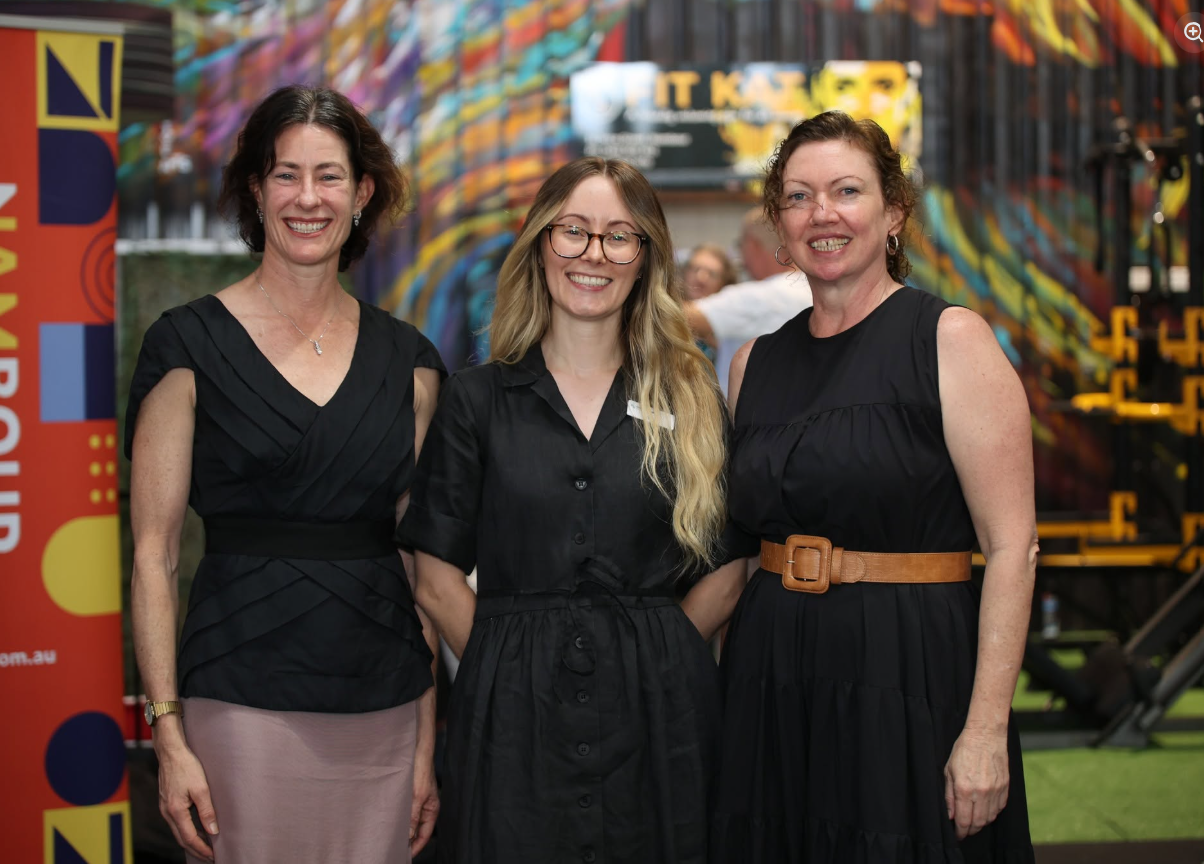 Three women standing together, smiling at an event with colorful wall art and a yellow and black playground structure in the background. Two women are wearing black dresses, and the woman in the middle has blonde hair and glasses.