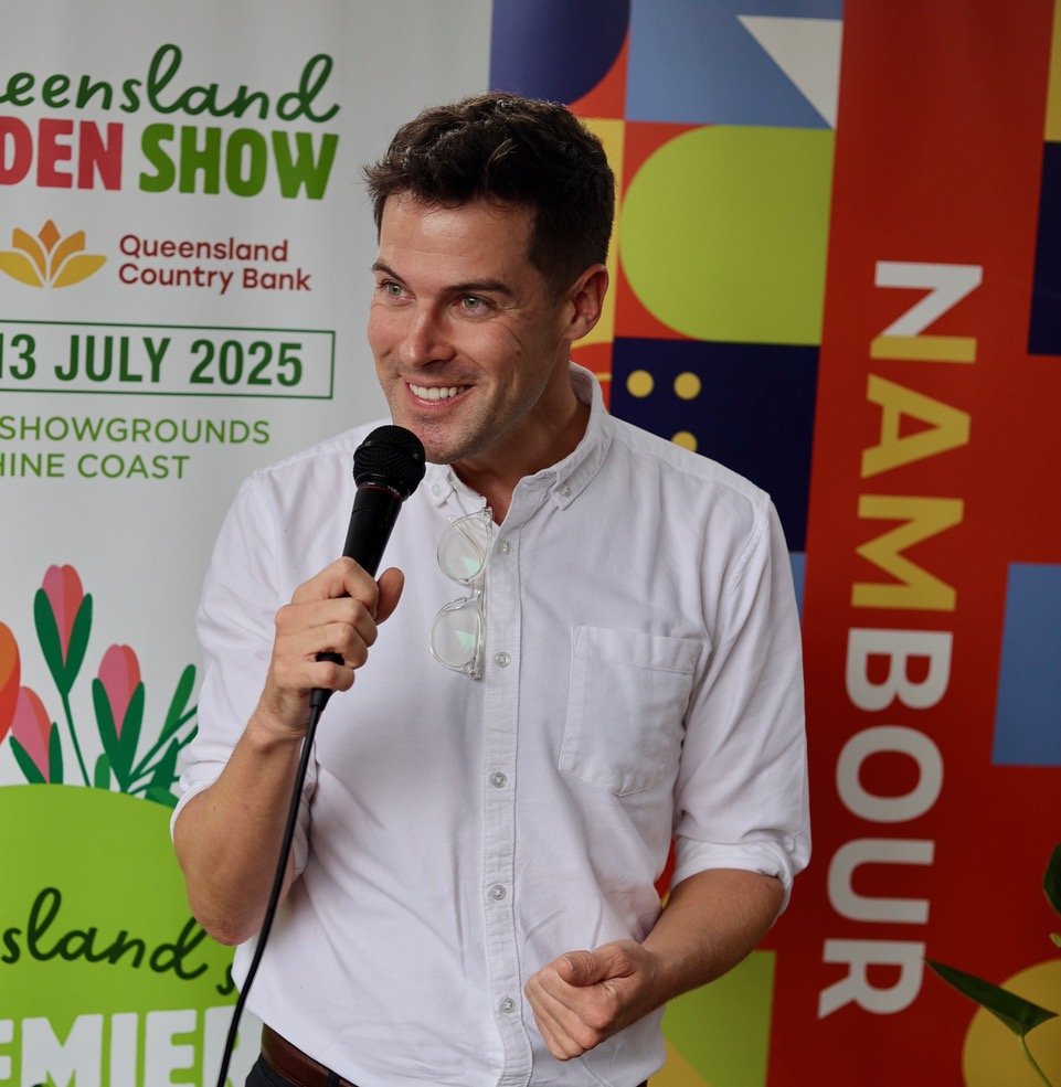 A smiling man holding a microphone at an event with colorful banners and posters in the background.