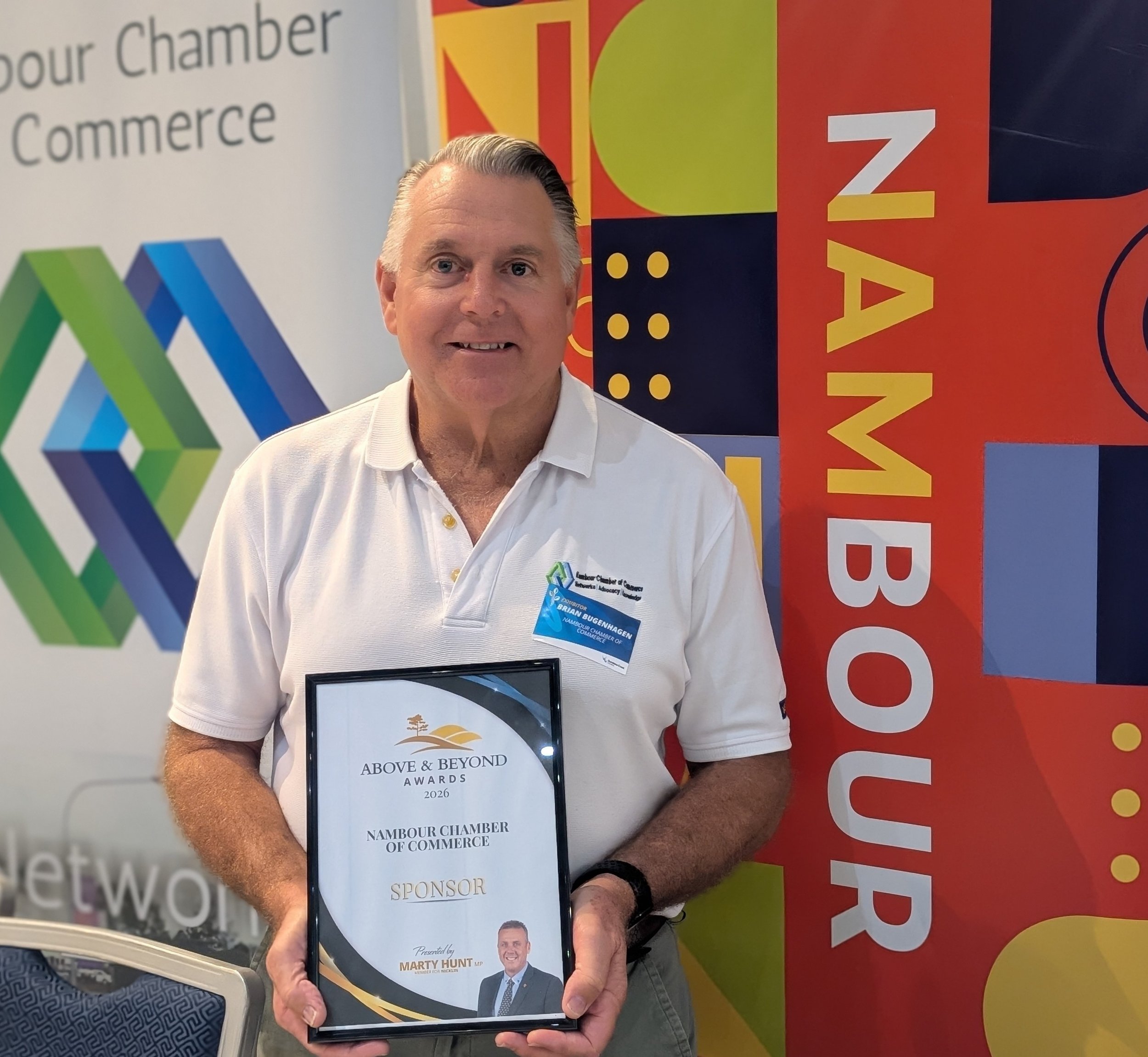 A man with gray hair holding an award plaque in front of a colorful backdrop with the words 'Nambuor Chamber of Commerce'. He is wearing a white polo shirt with a name badge that reads 'Brian Buggenhagen'. The plaque reads 'Above & Beyond Awards 2026