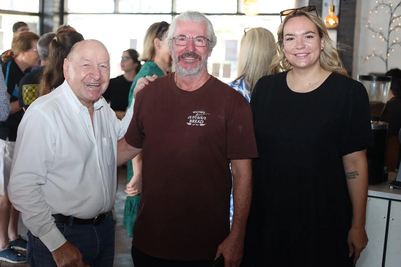 Three people smiling at a social gathering inside, with many people in the background, including a man with glasses and a beard wearing a brown t-shirt, an elderly man in a white shirt, and a young woman with blonde hair in a black shirt.