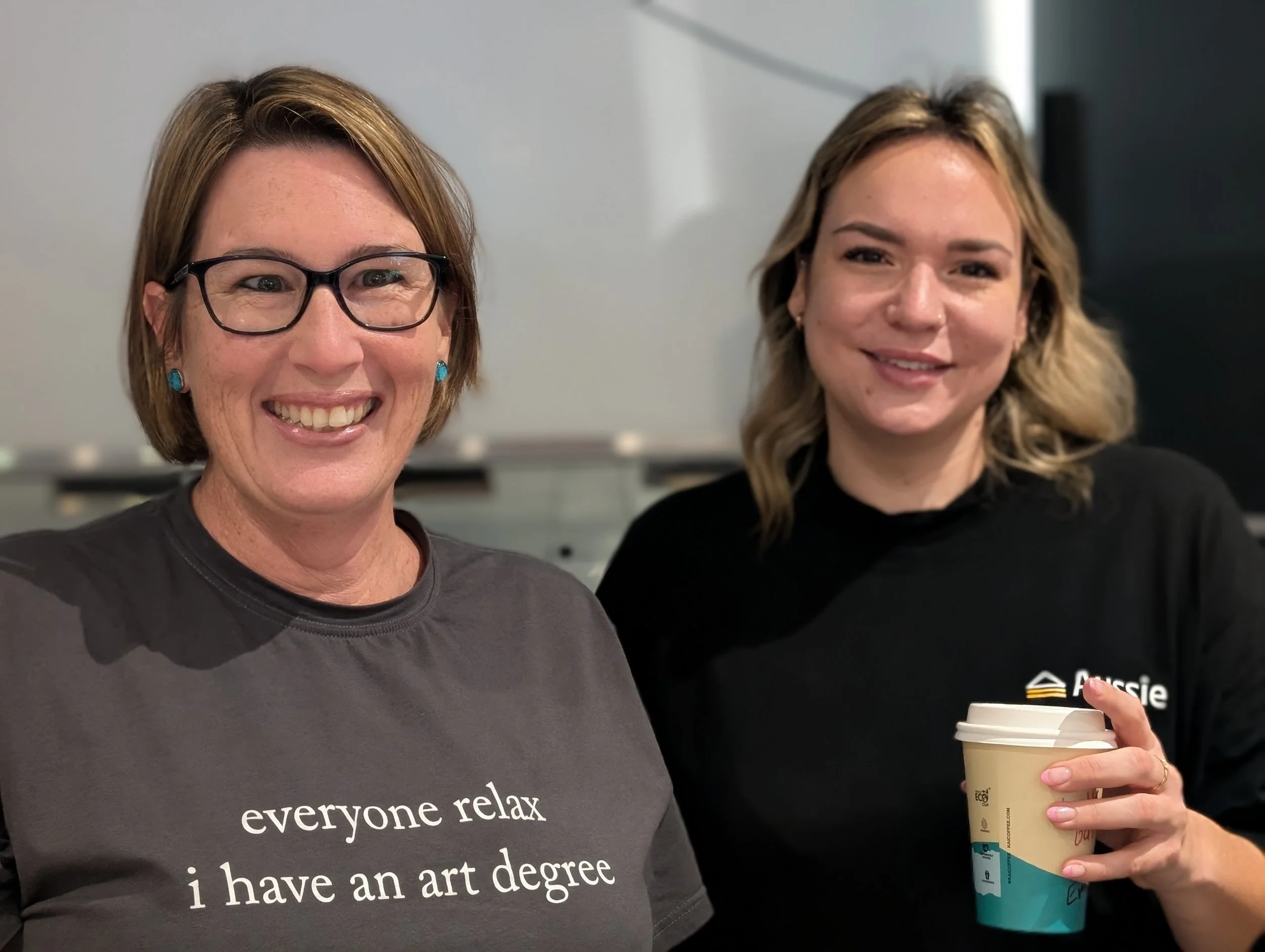 Two women standing close together, smiling. The woman on the left has short brown hair, glasses, and is wearing a gray shirt with white text. The woman on the right has wavy blonde hair and is holding a disposable coffee cup.