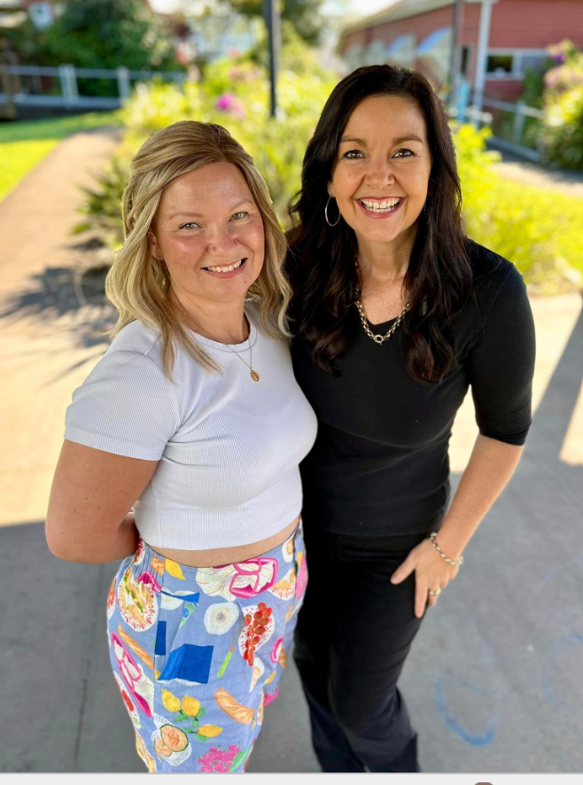 Two women smiling outdoors with a garden and a house in the background, one wearing a white crop top and colorful patterned pants, the other wearing a black top and black pants.