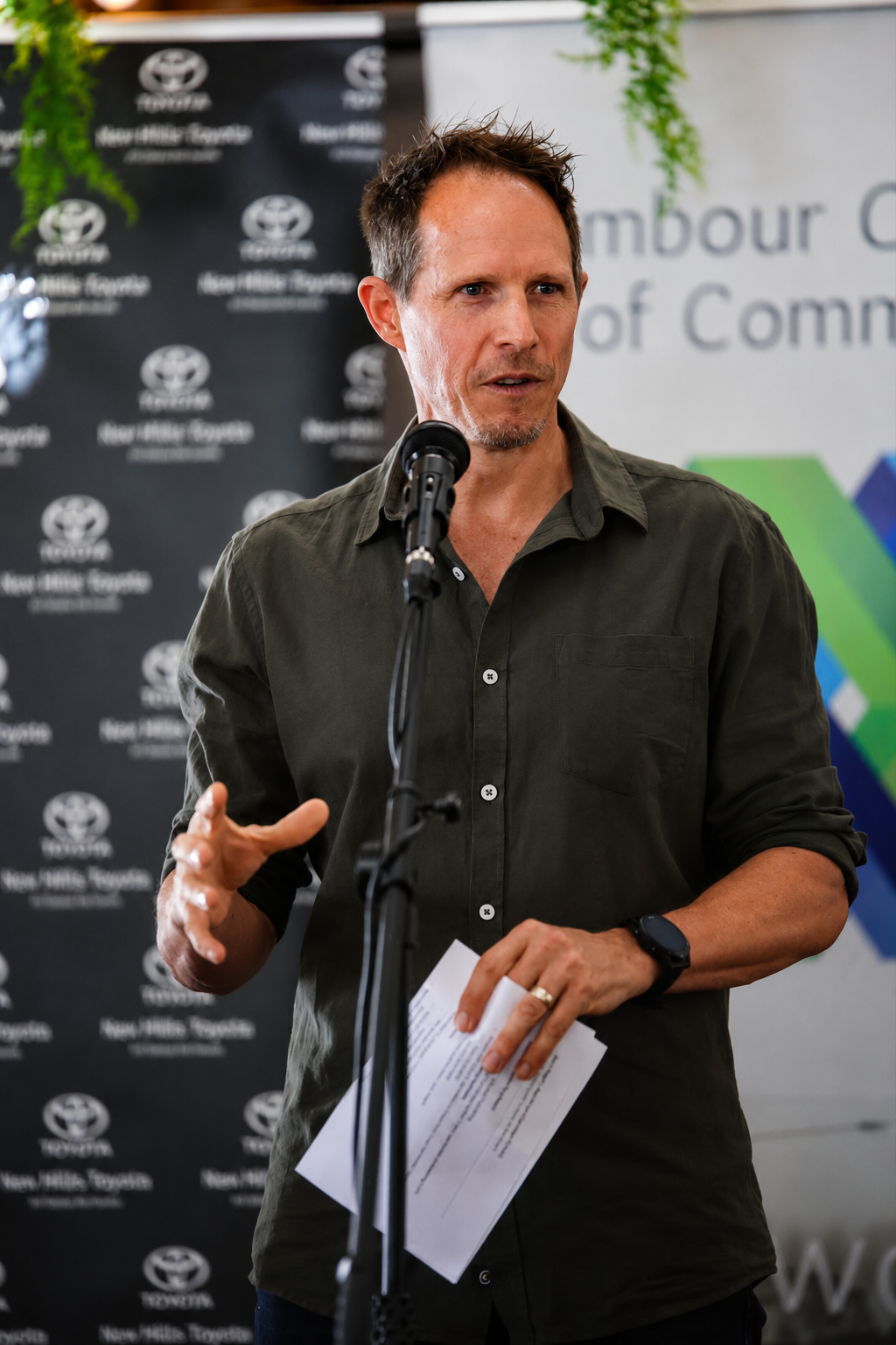 A man in a dark green button-up shirt speaking into a microphone at a press conference, holding papers, with a backdrop displaying the New Hills Toyota logo.