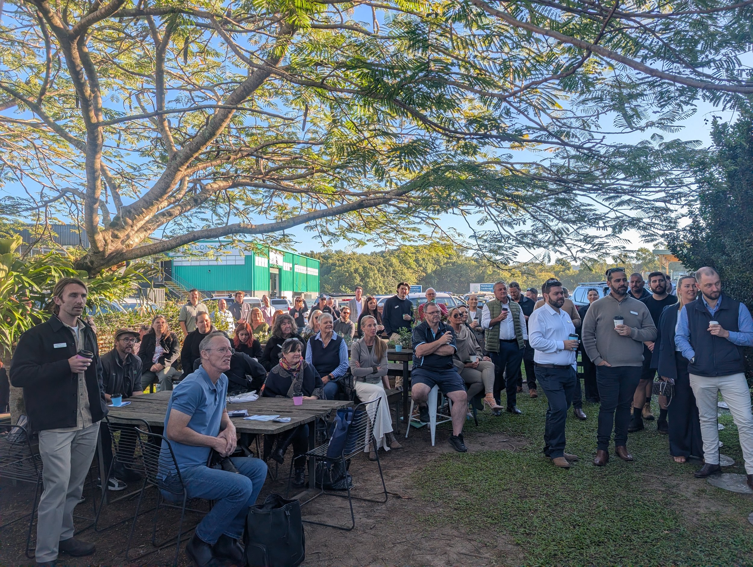 A diverse group of people gathered outdoors under a large tree, listening attentively to a speaker at an event on a sunny day.