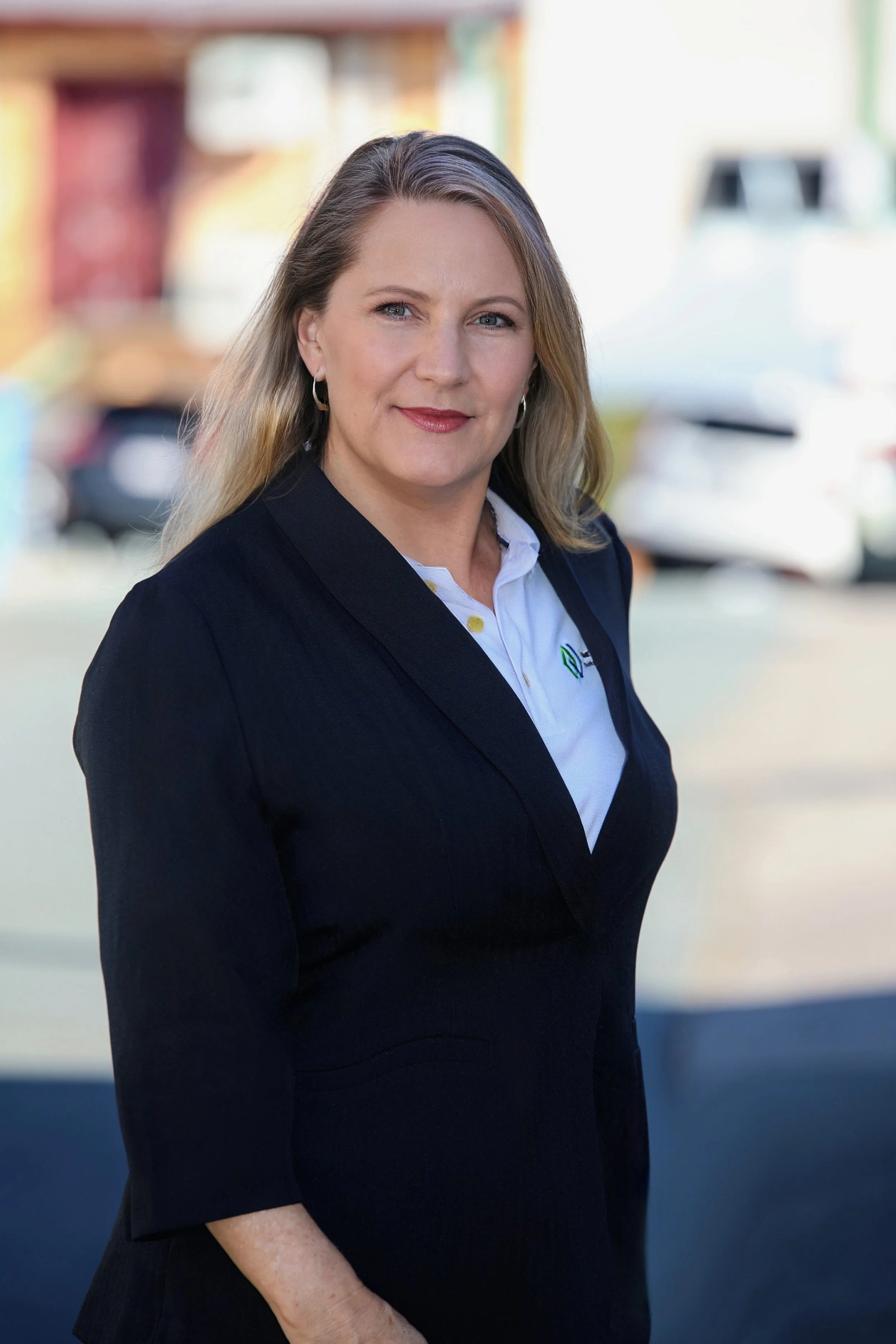A middle-aged woman with blonde hair, wearing a black blazer over a white shirt, standing outdoors with blurred background of buildings and cars.