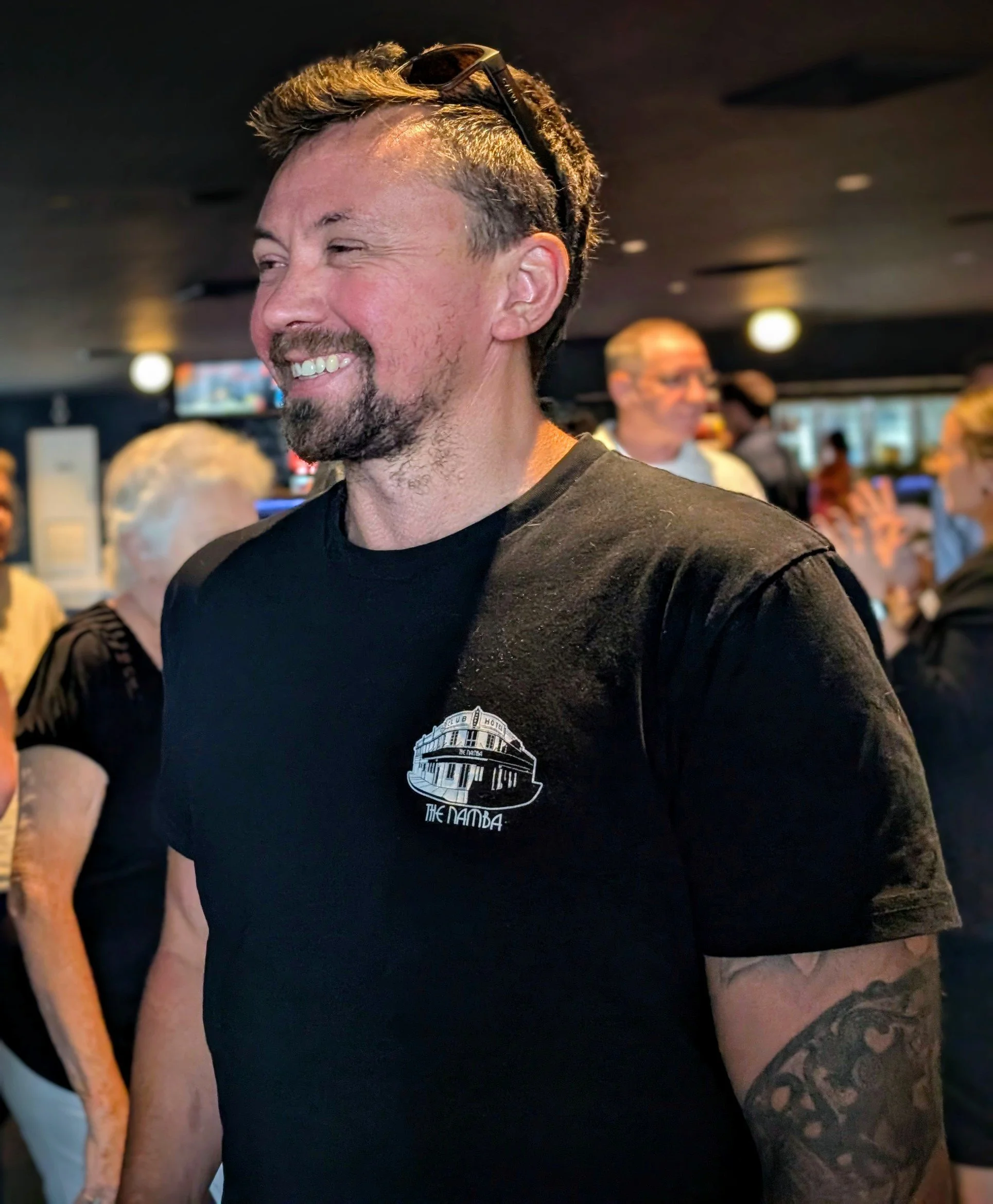 Close-up of a smiling man with sunglasses on his head, wearing a black t-shirt with a graphic of a building and the text 'The Namba', standing in what appears to be a busy indoor setting, possibly a bar or restaurant.