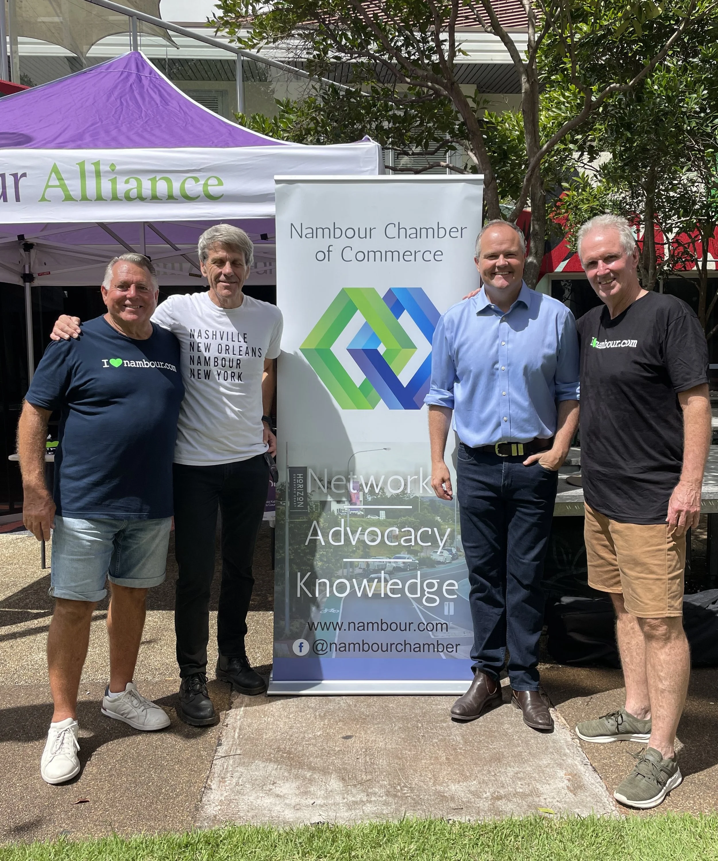 Four men smiling outdoors near a banner and a purple canopy tent. They are standing on a sidewalk with greenery at their feet. The banner displays 'Nambour Chamber of Commerce' with a colorful geometric logo and the words 'Network Advocacy Knowledge'