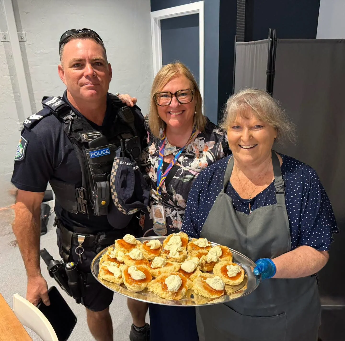 Three women and one police officer smiling; woman on right holding a tray of appetizers topped with cream cheese and jam.