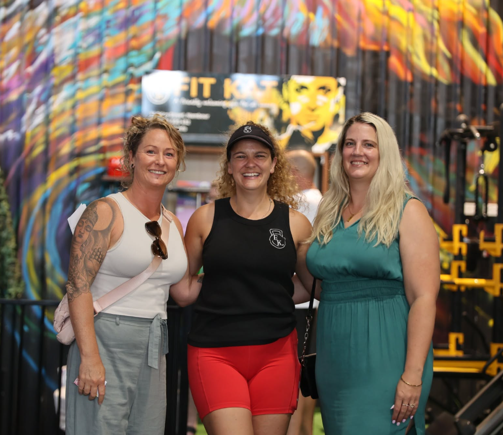 Three women smiling and posing together indoors at a colorful gym with climbing walls in the background.