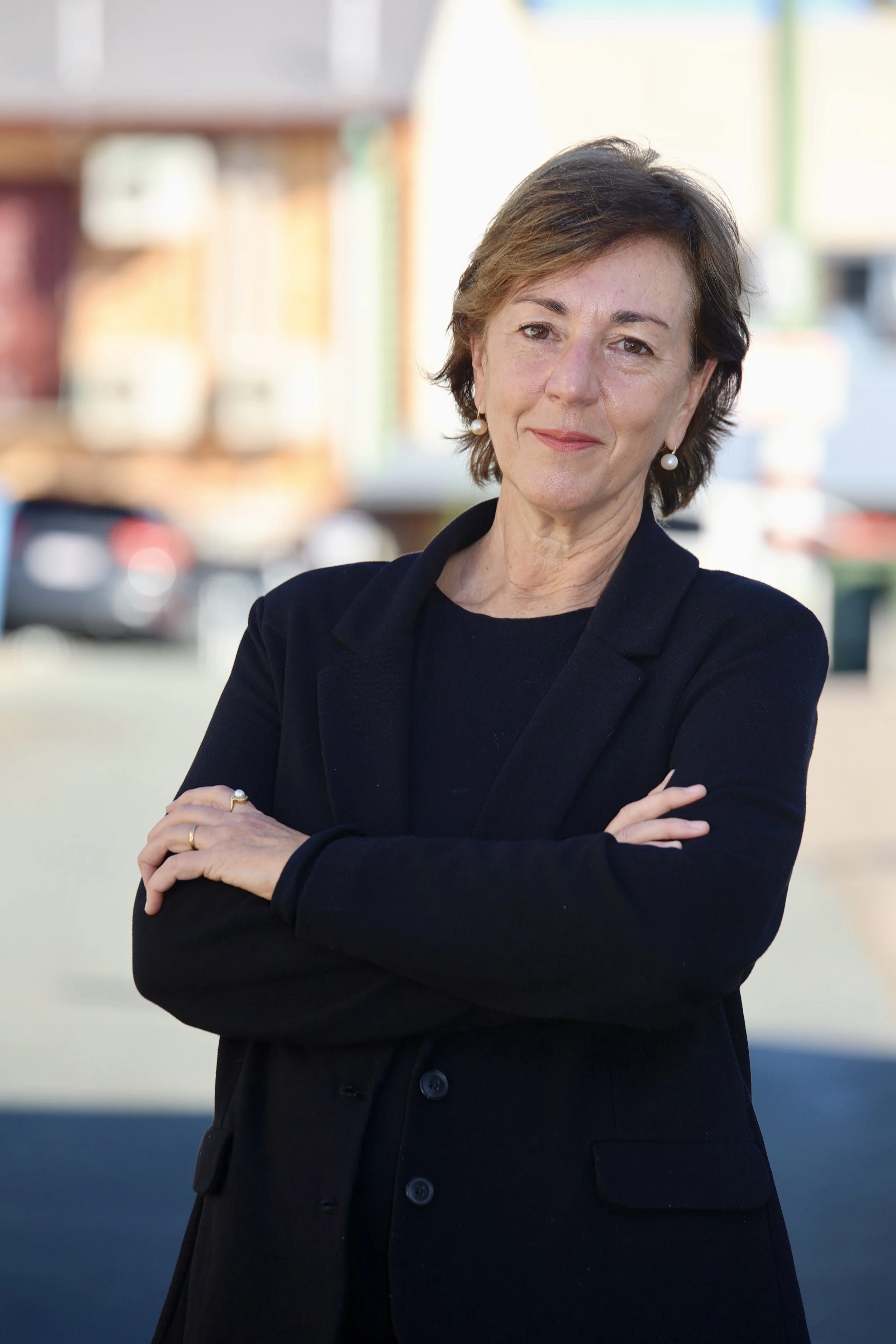 A woman with short brown hair wearing pearl earrings and a black jacket standing outdoors with her arms crossed, smiling subtly, with a blurred background of a parking lot and buildings.
