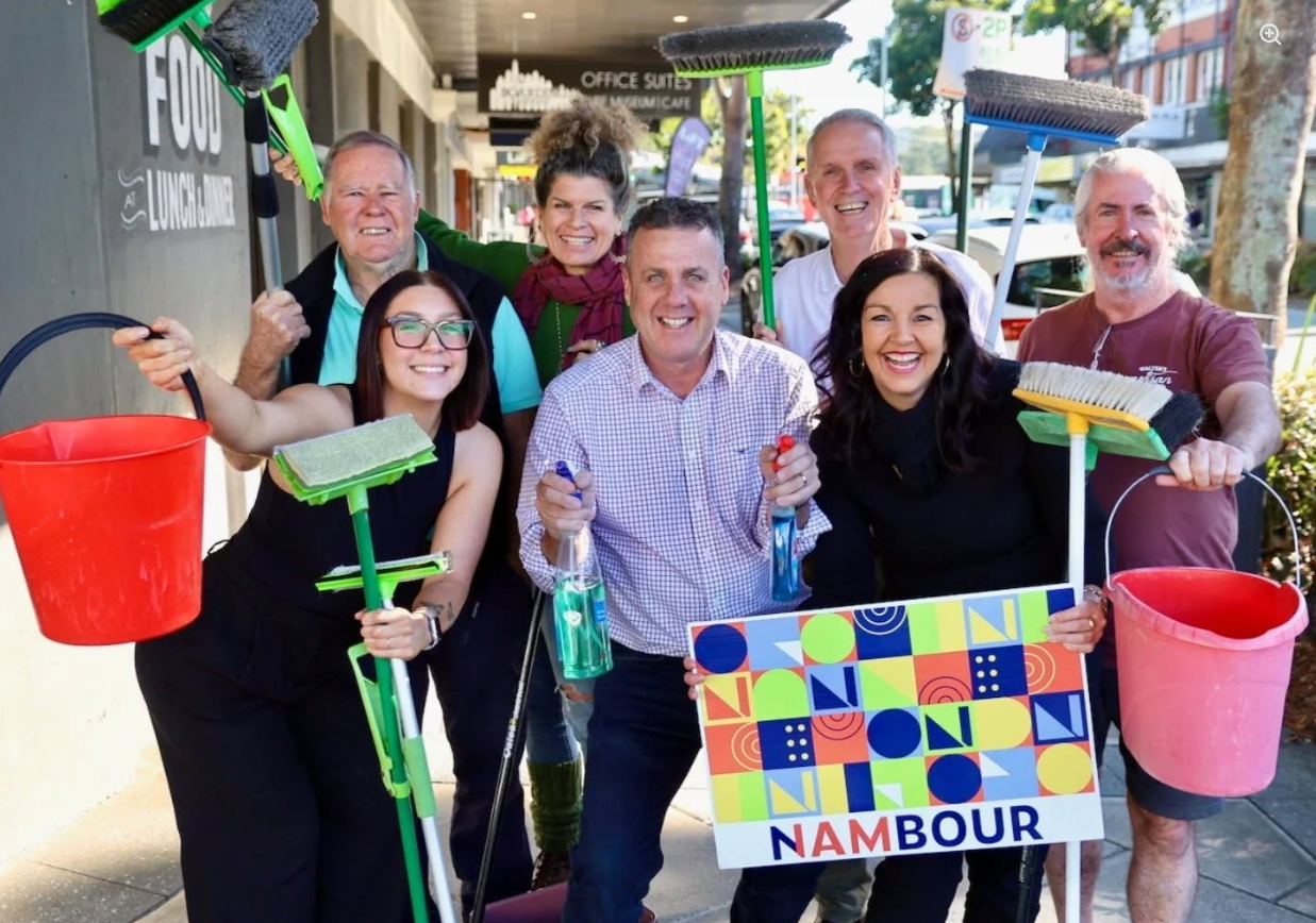 Group of eight smiling people holding cleaning tools and spray bottles, standing on a sidewalk next to a colorful sign that says 'Nambour'. They appear cheerful and ready for community cleanup.