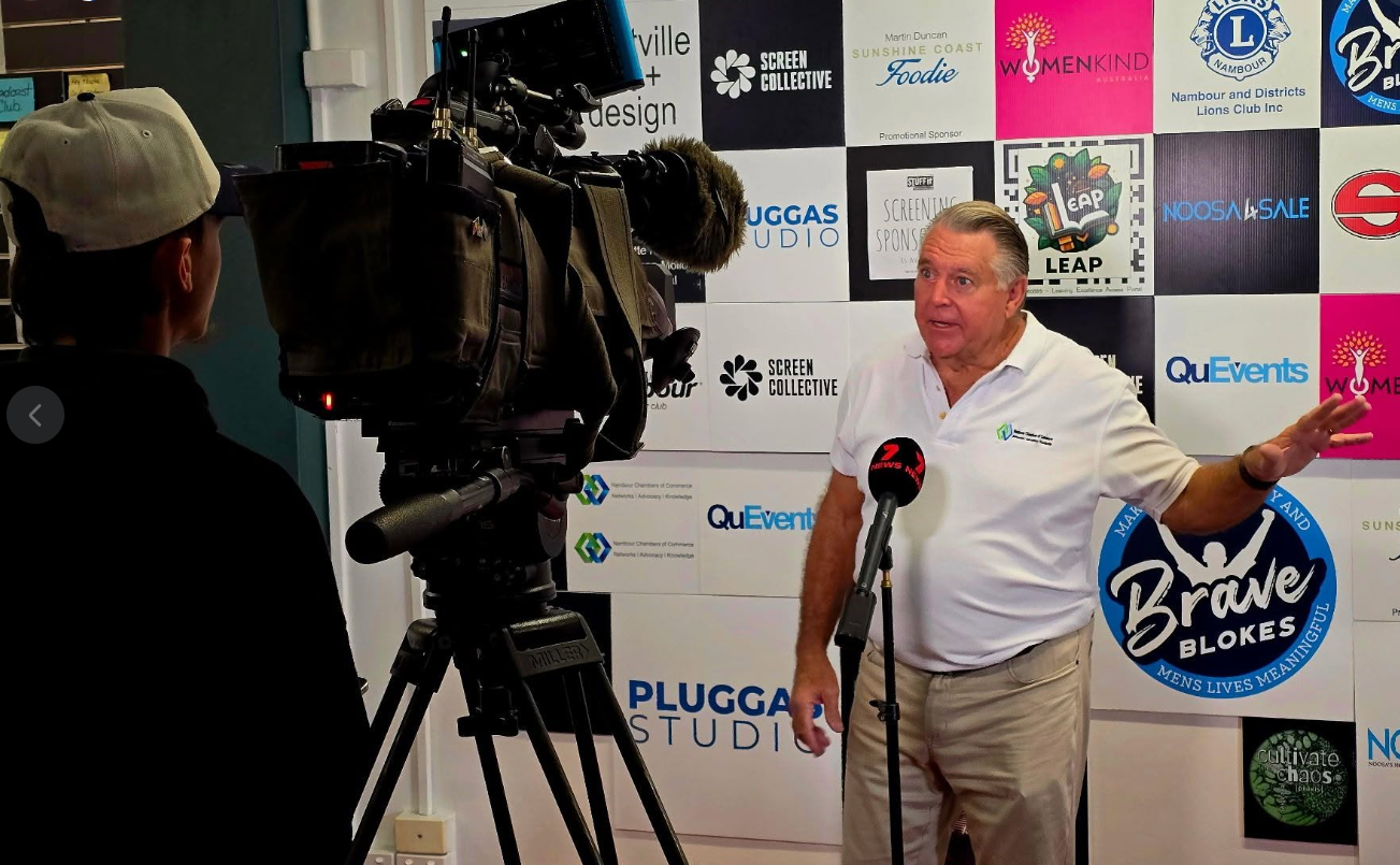 A man wearing a white polo shirt speaking into a microphone during an interview or press conference with a backdrop featuring multiple logos and signs.