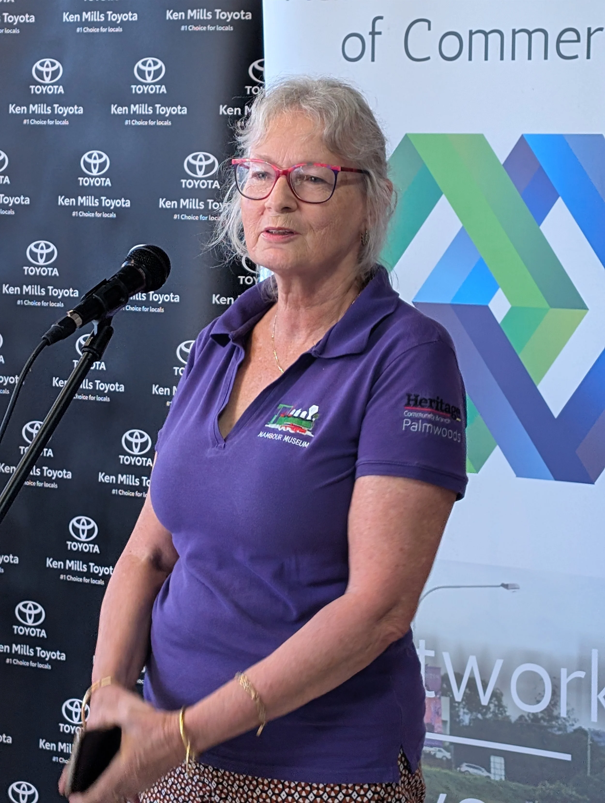 A woman with glasses and gray hair speaking into a microphone, wearing a purple polo shirt with logos, standing in front of a backdrop with Toyota and Ken Mills Toyota logos, and a geometric patterned banner.