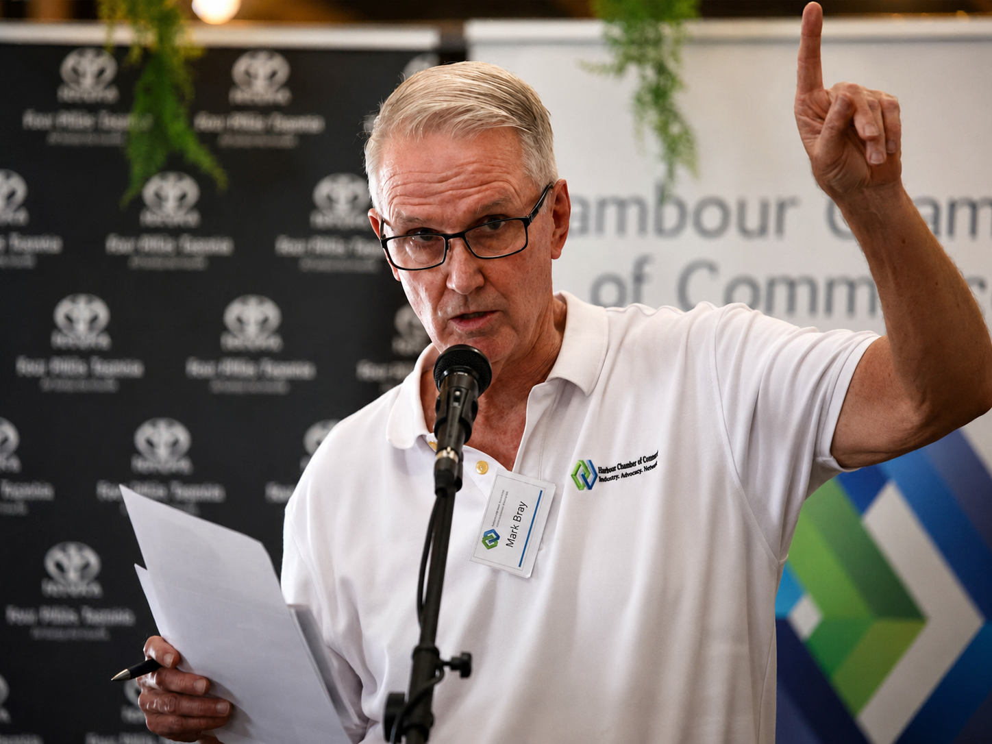 A man with gray hair and glasses speaking into a microphone while holding papers, raising his right index finger, wearing a white polo shirt with a name tag that reads 'Mark Bray', standing in front of a black backdrop with logos and a banner for the Bar Harbor Chamber of Commerce.