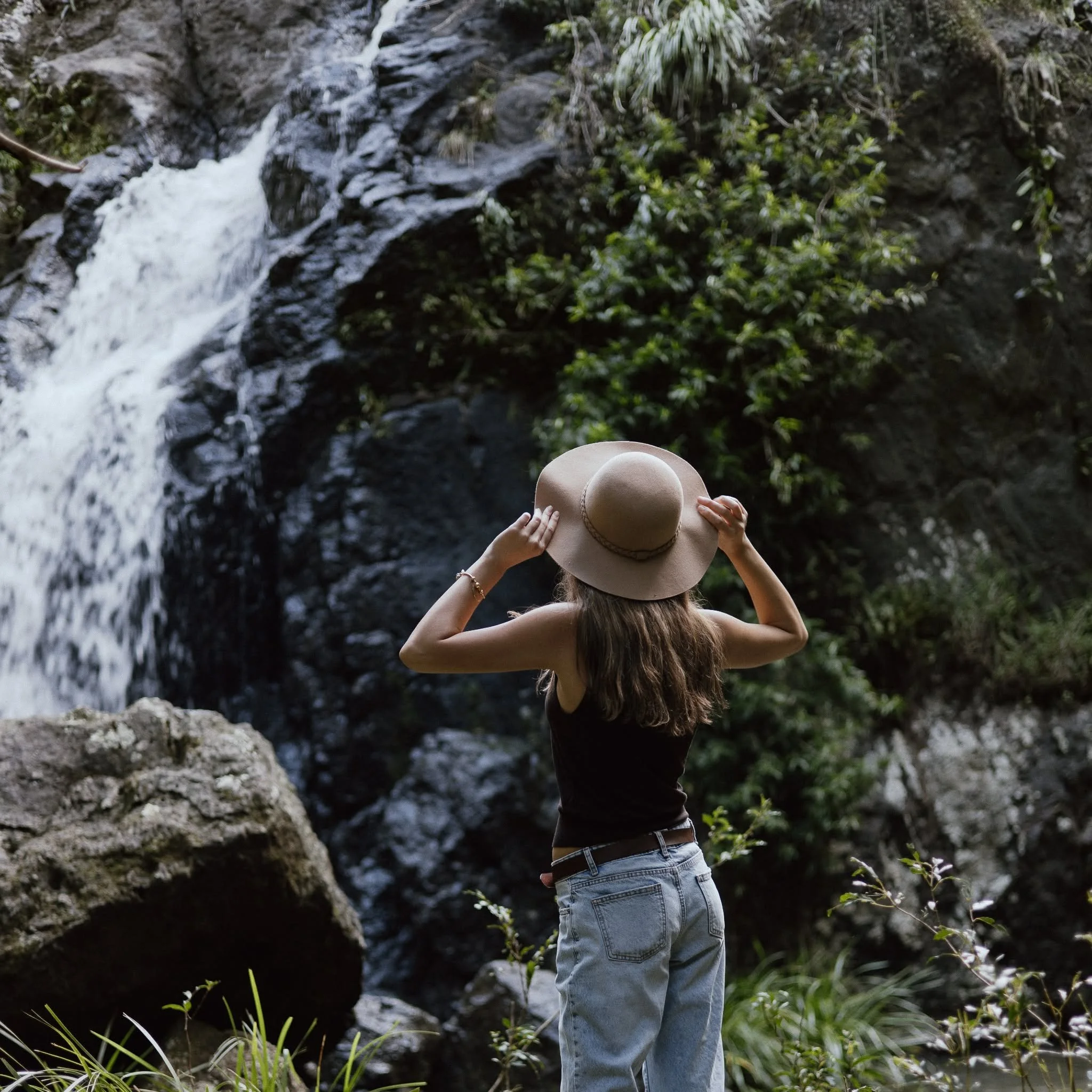 A woman with long hair wearing a straw hat, black tank top, and jeans standing near a waterfall surrounded by greenery, viewed from behind.