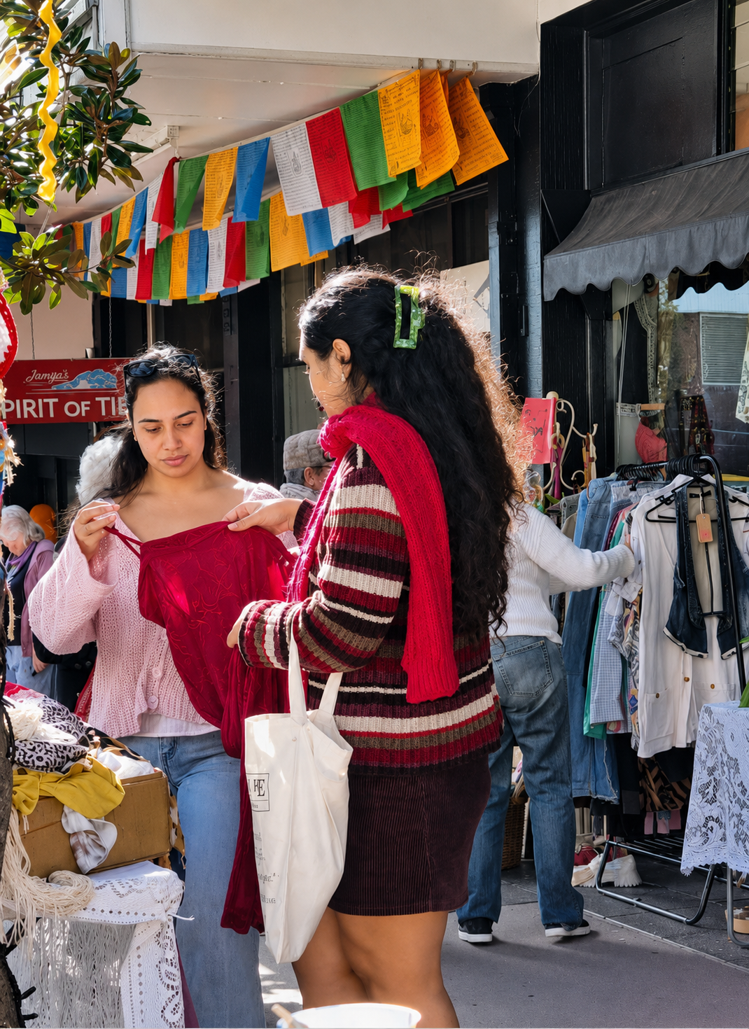 Two women shopping at an outdoor market, one holding a red garment and the other holding a white tote bag, with clothes displayed on racks and tables around them.