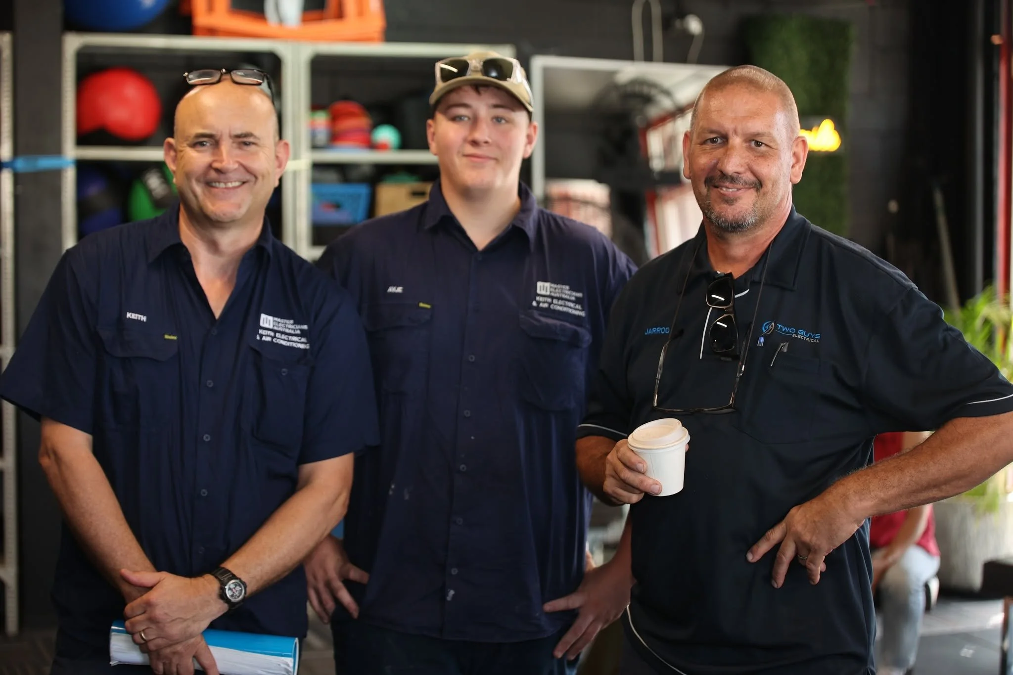 Three men standing together in an indoor space with shelves of equipment in the background. They are smiling and wearing work shirts with company logos. One man on the right is holding a coffee cup.