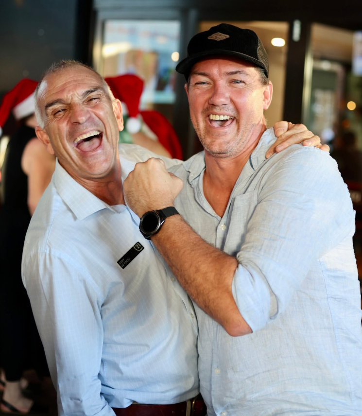 Two men smiling and laughing together, one wearing a white shirt and the other in a light gray shirt and a black baseball cap, in a festive setting with holiday decorations in the background.
