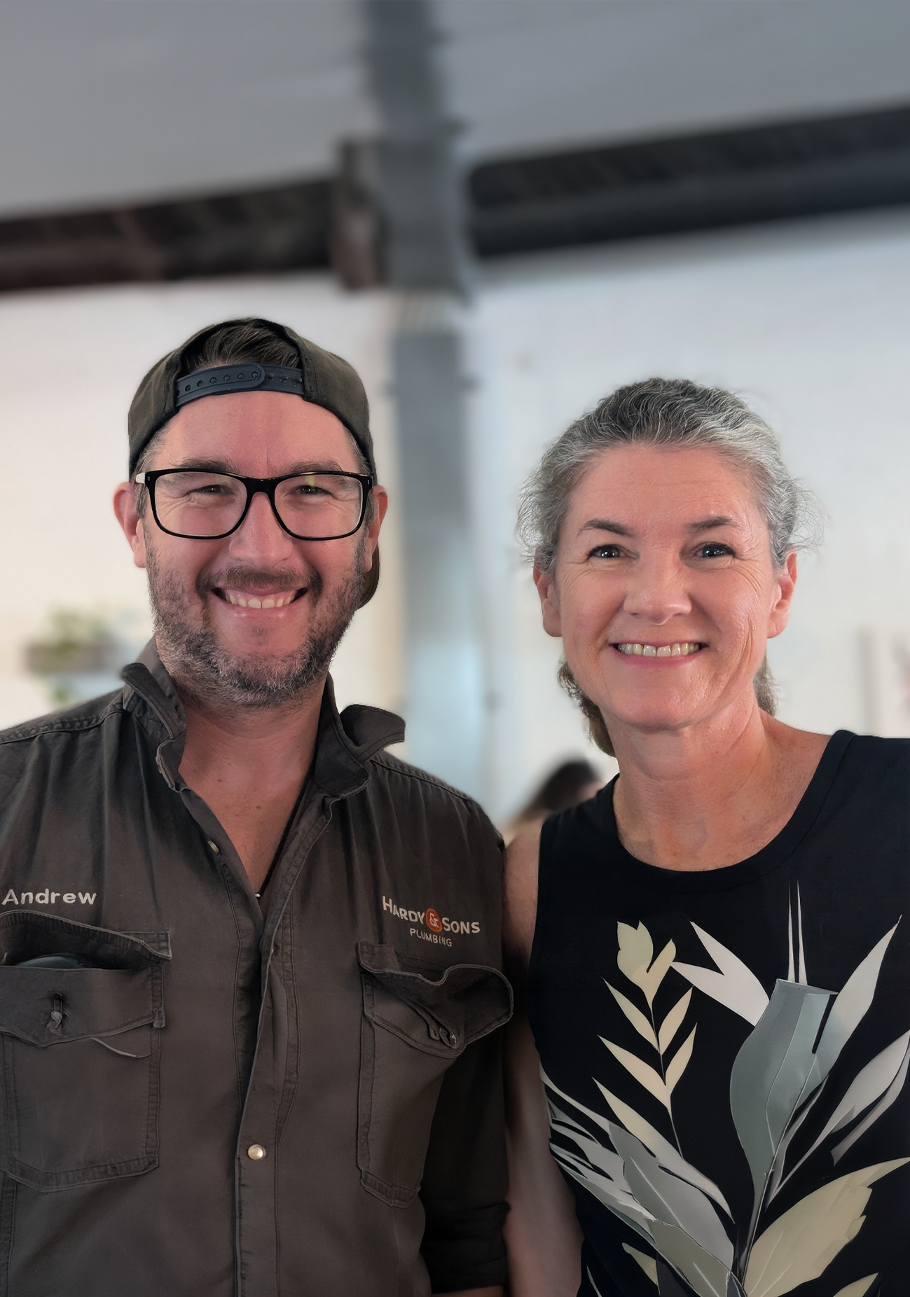 A smiling man wearing glasses, a backward baseball cap, and a gray work shirt with the name tag 'Andrew' and a company logo, standing next to a smiling woman with gray hair wearing a black top with a white floral pattern.