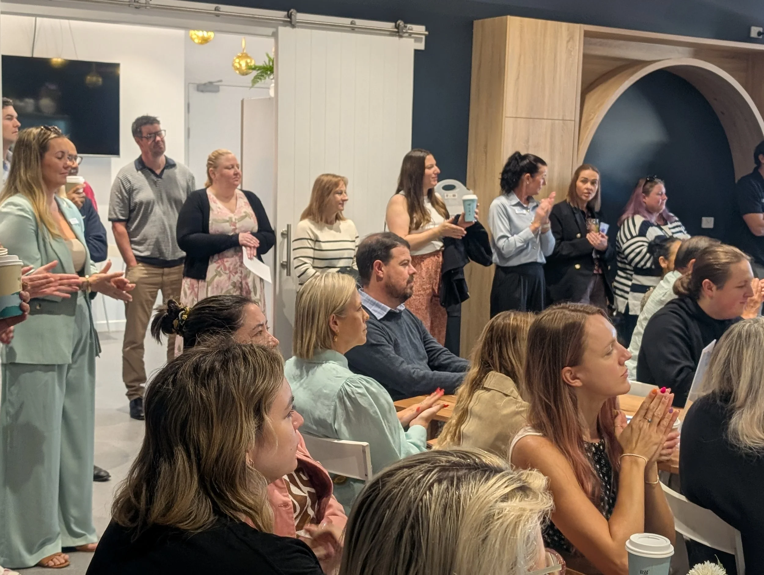 A group of people standing and sitting in an indoor meeting or conference room, listening attentively, with some holding coffee cups.