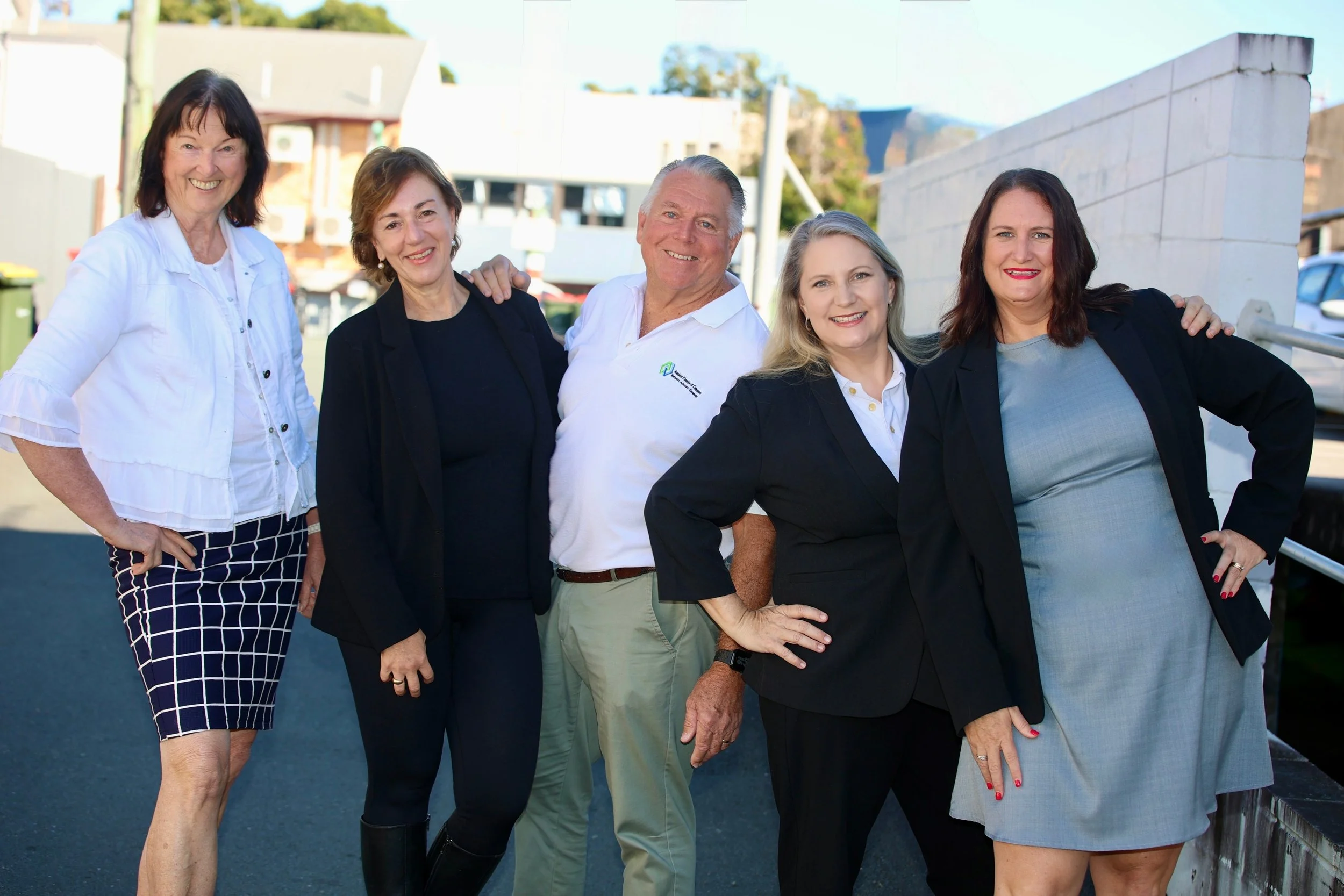 A group of five diverse adults, three women and two men, smiling outdoors, dressed in business casual attire, standing together with their arms around each other near a white wall and buildings.