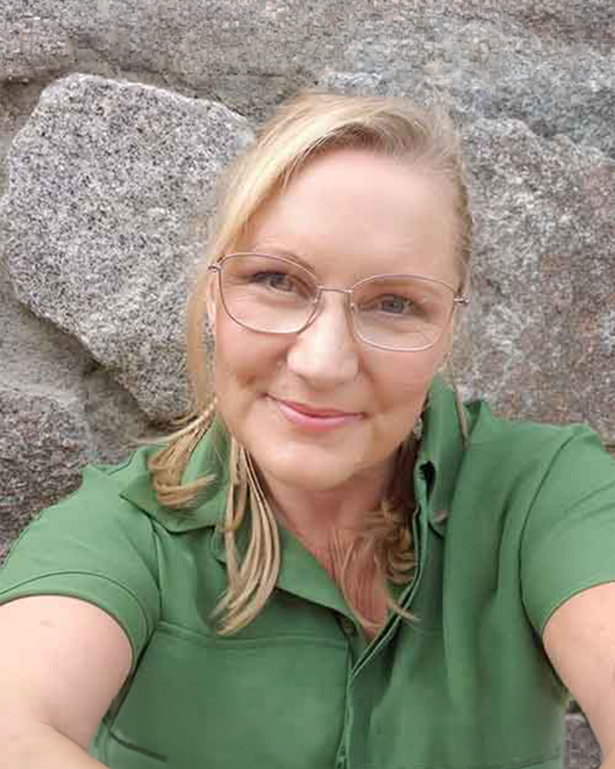 A woman with blonde hair and glasses smiling for a selfie against a rocky background, wearing a green polo shirt.
