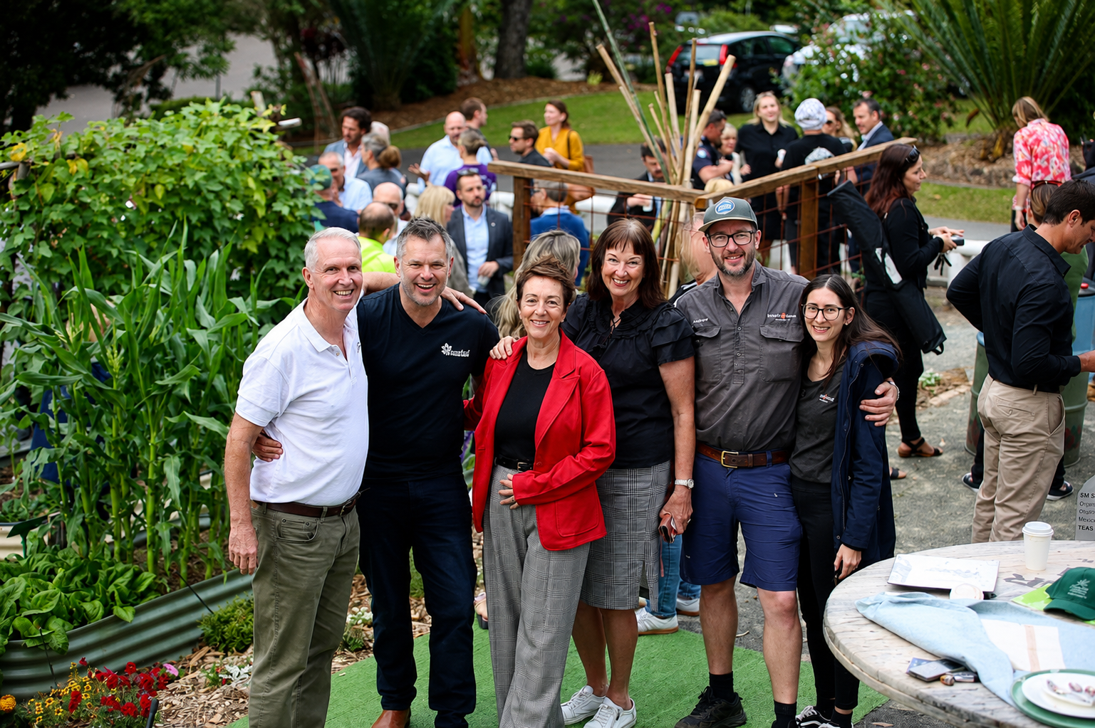 Group of six people smiling and posing together outdoors at a garden event, with more people in the background and garden plants nearby.