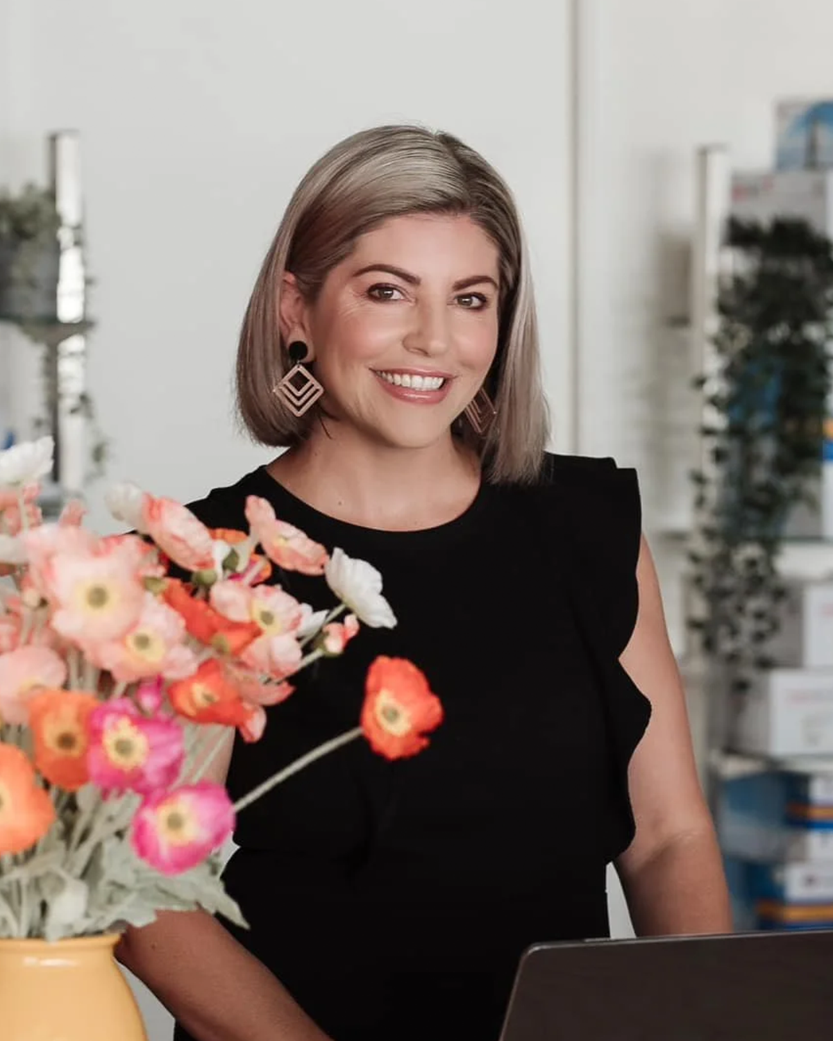 A woman with shoulder-length gray hair, wearing a black sleeveless top and geometric earrings, smiling while standing in a bright office with shelves and boxes in the background. A bouquet of pink, orange, and white flowers is in the foreground.