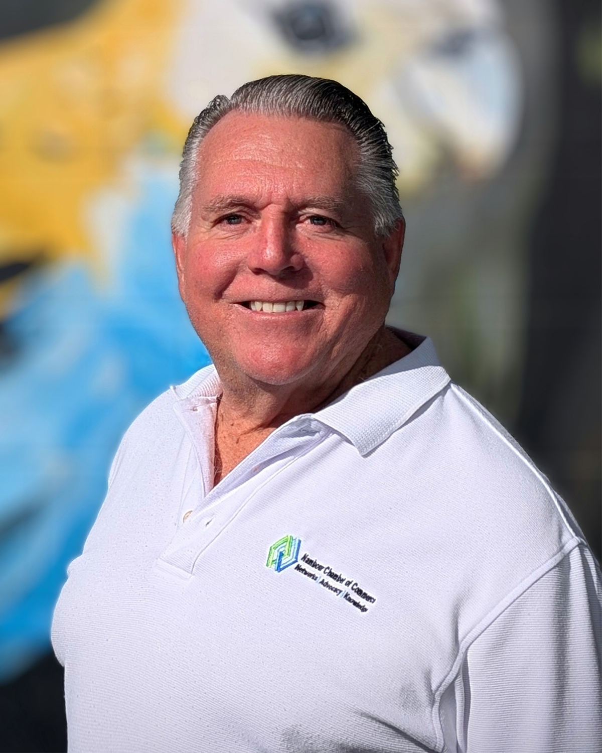 A smiling man in a white polo shirt with a logo that reads 'Northeast Chamber of Commerce, Networking, Advocacy, Community' stands outdoors in front of a blurred colorful background.